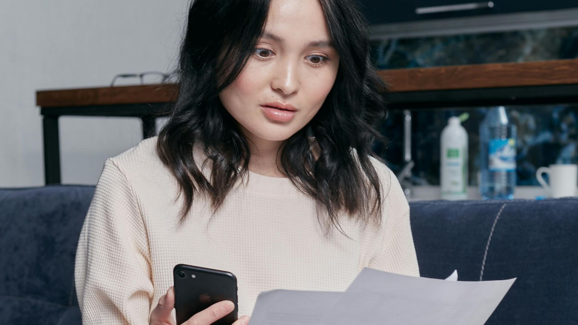 Young woman reviews documents and phone indoors, showcasing concern while seated on a couch with scattered papers. Business concept.