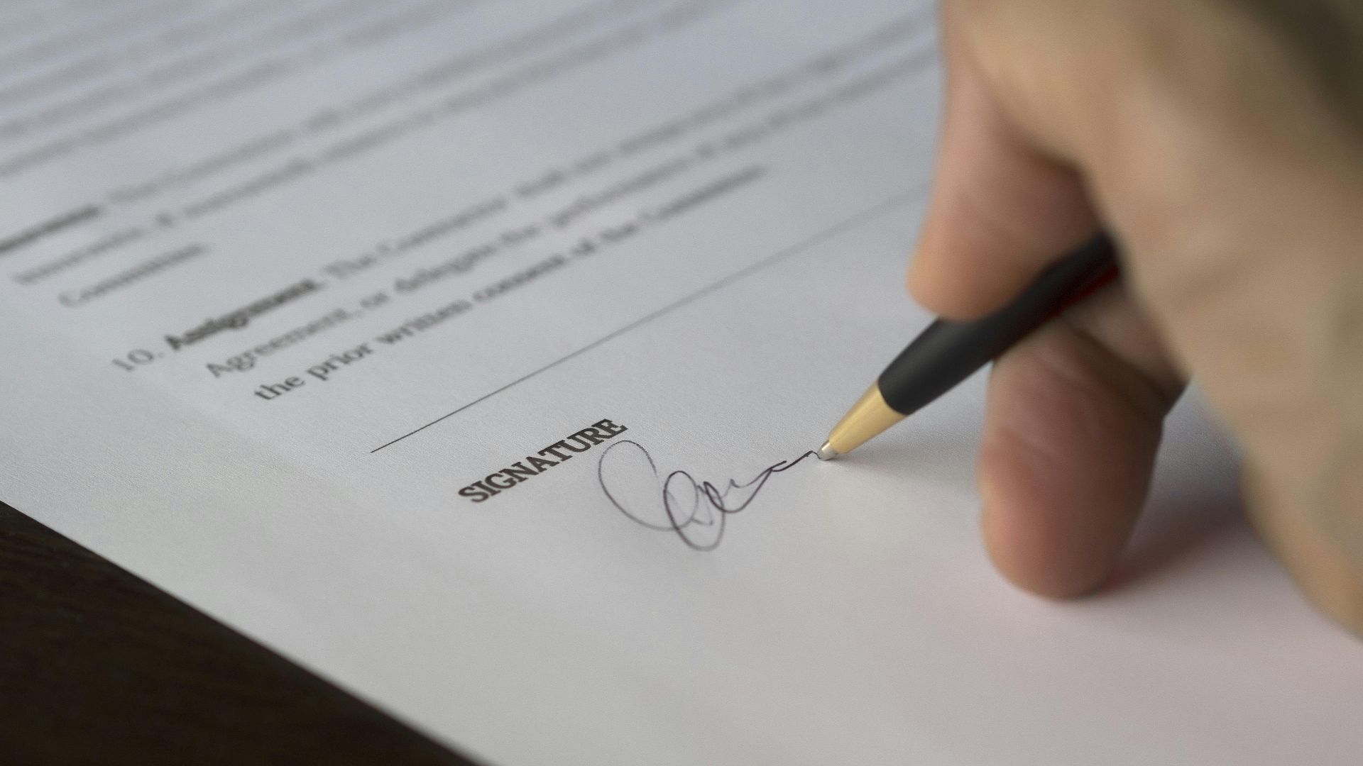 A hand signs a formal contract with a pen on a wooden desk.