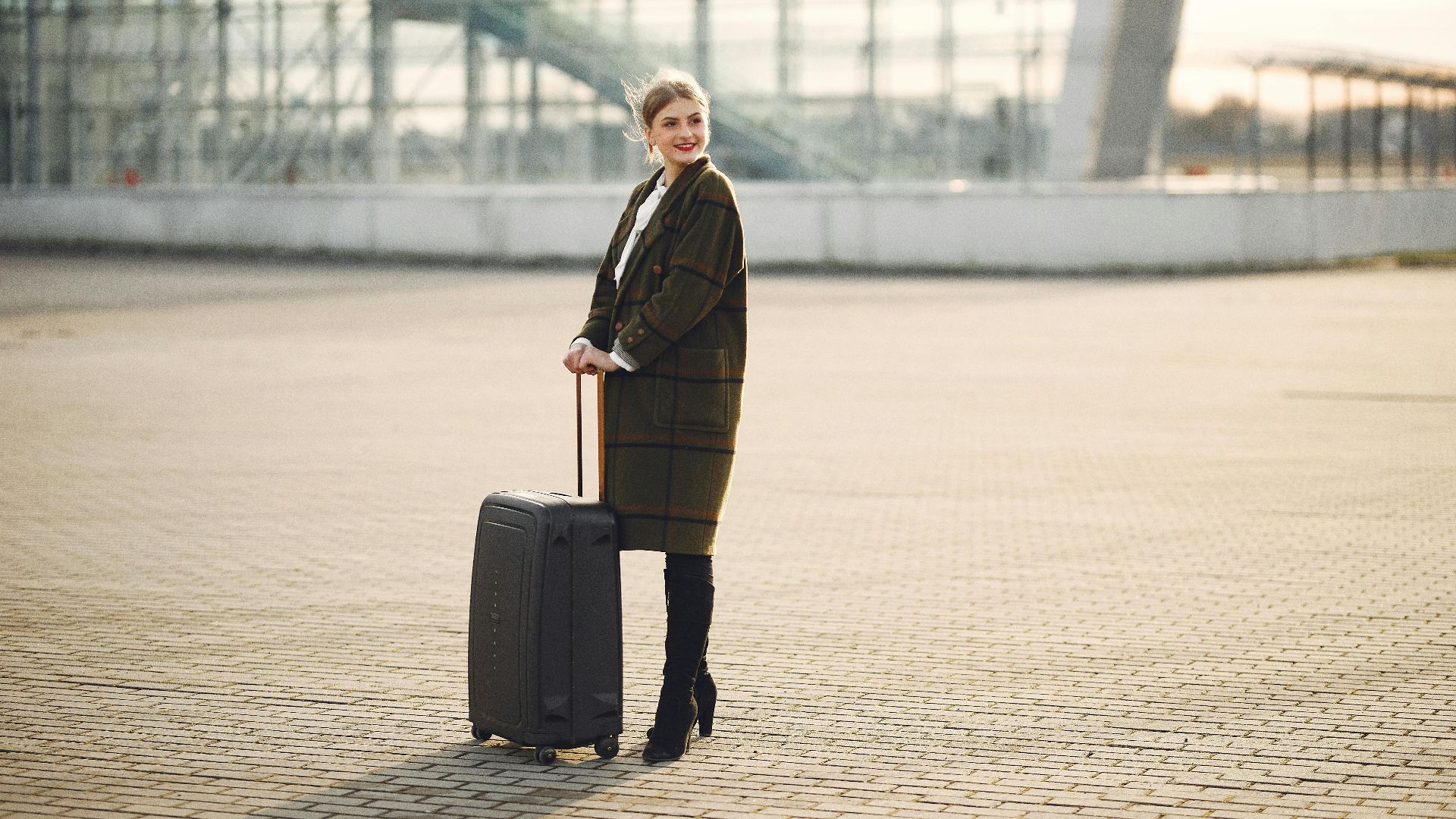 Full length of young joyful woman in outwear and black high heeled boots with luggage standing against glass wall of airport building and looking away