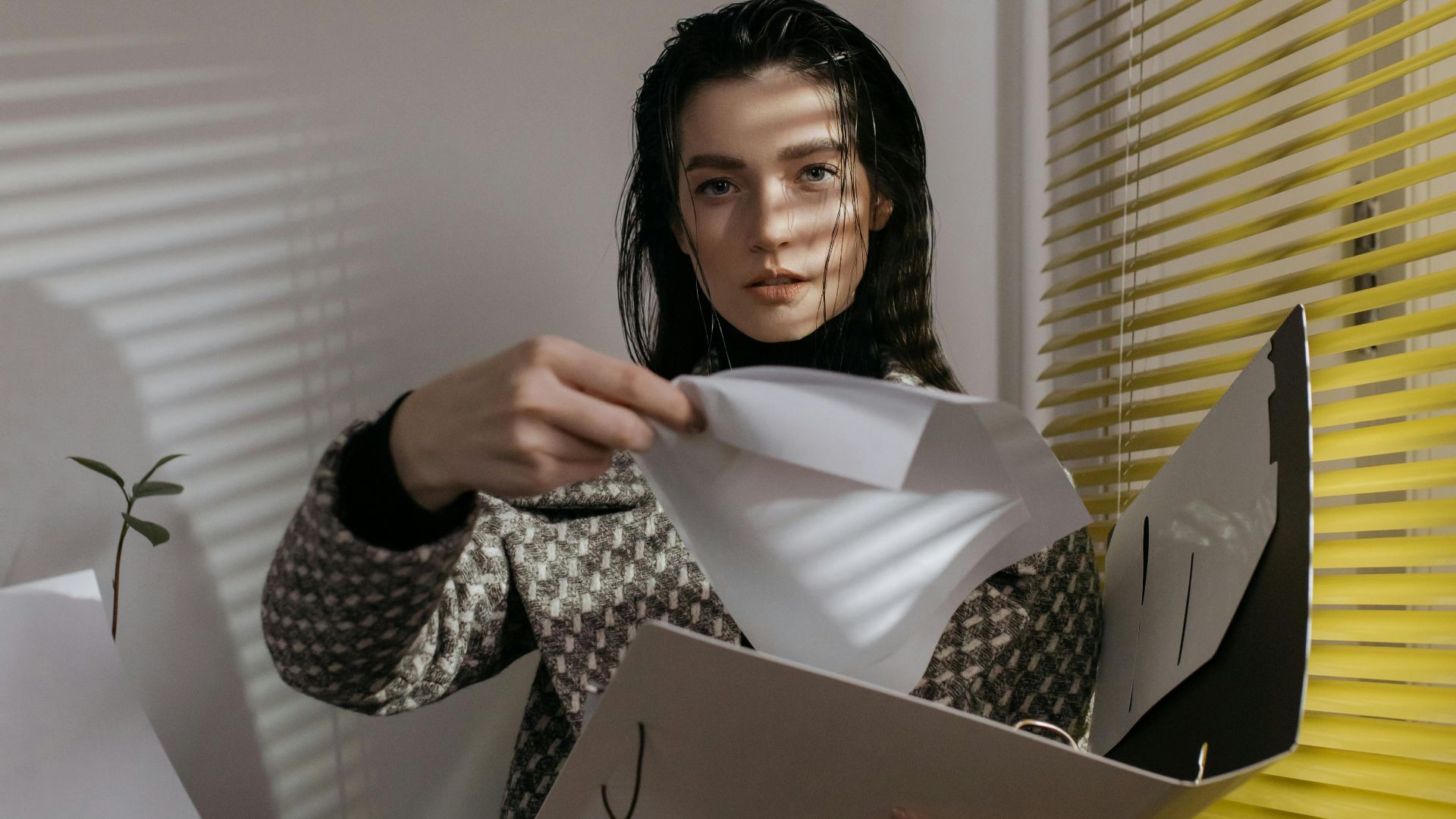 A woman is sorting papers in a modern office, with light filtering through blinds.