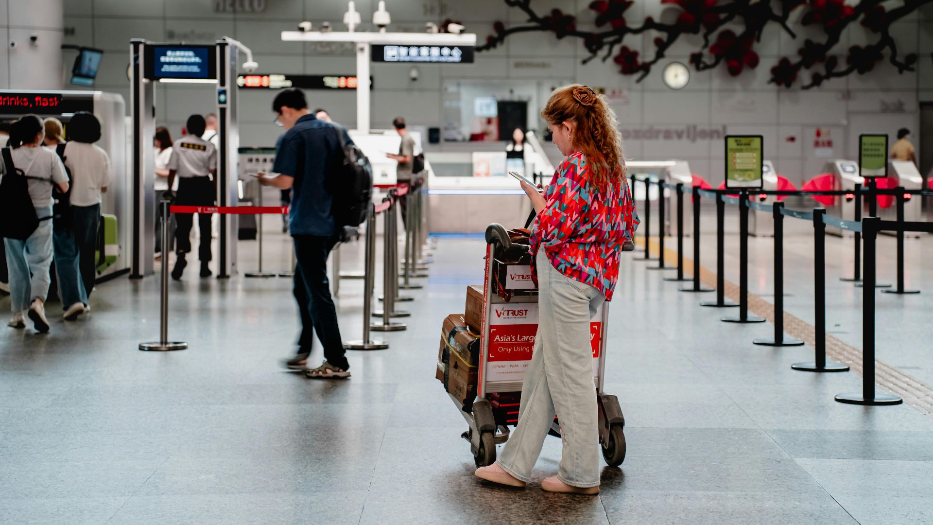 A traveler checks her phone while waiting with a luggage cart at a busy airport terminal in Guangzhou.