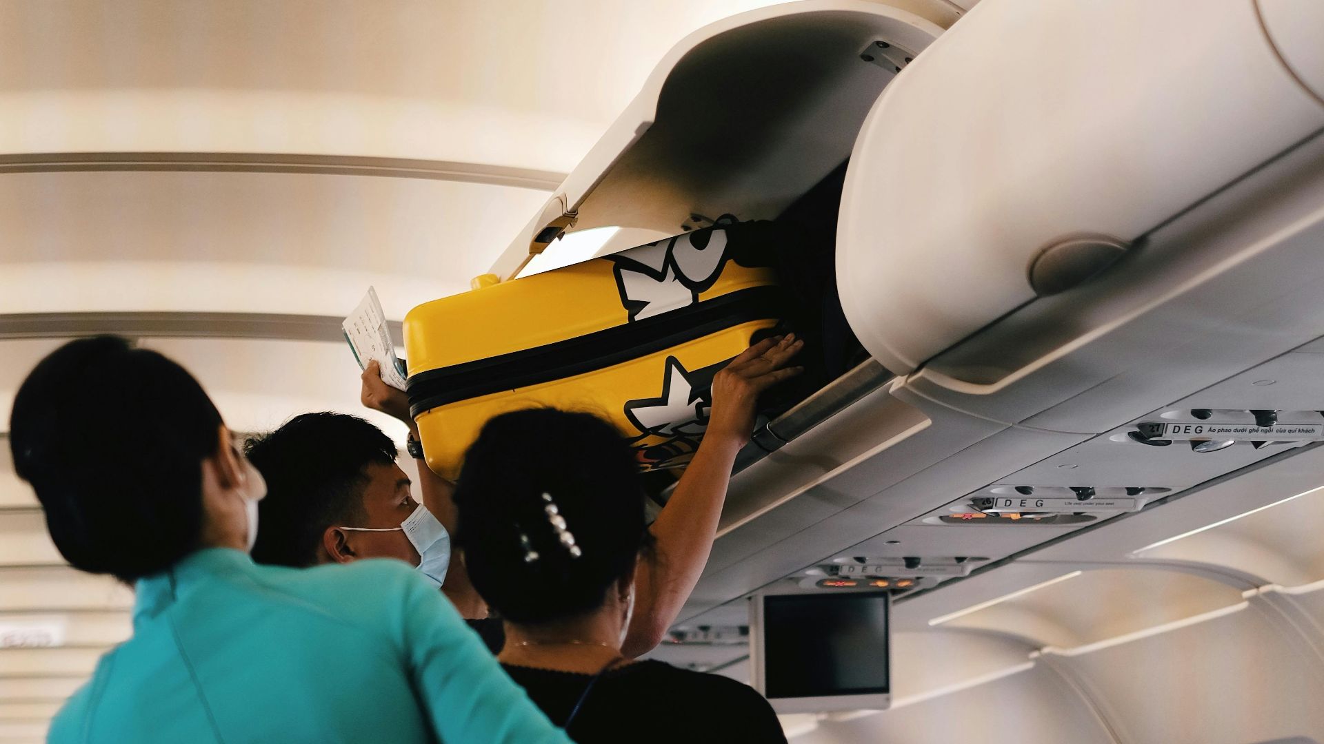 Passengers placing a yellow suitcase in the overhead bin of an airplane cabin. Interior with crew assistance.