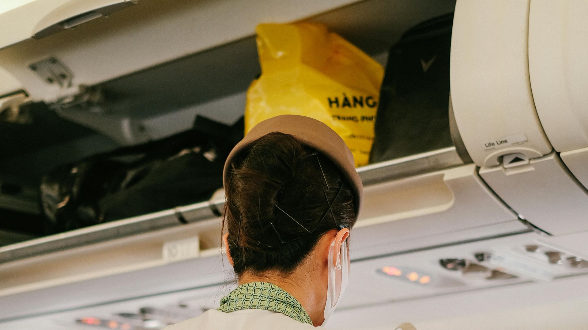 A flight attendant assists passengers with luggage in an airplane cabin.