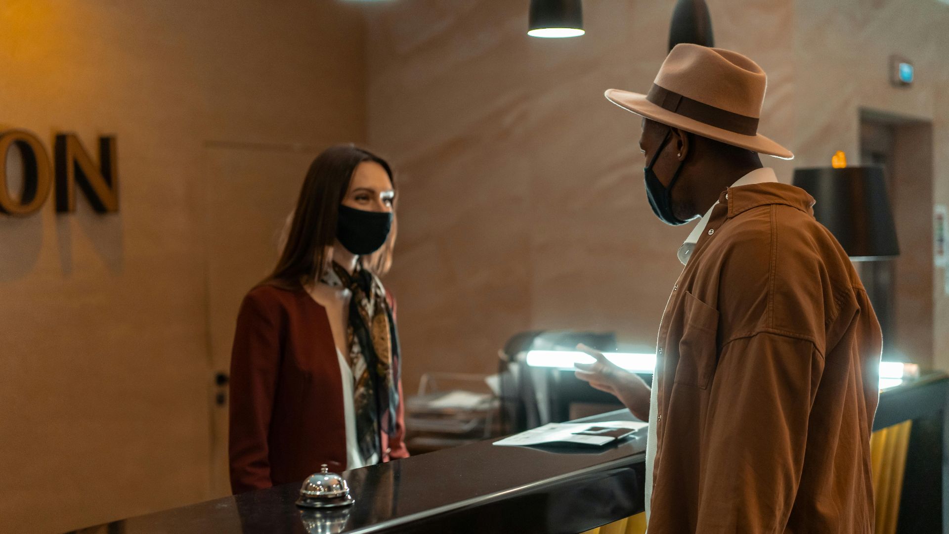 A man at a hotel reception talking to a staff member. Both wear face masks.