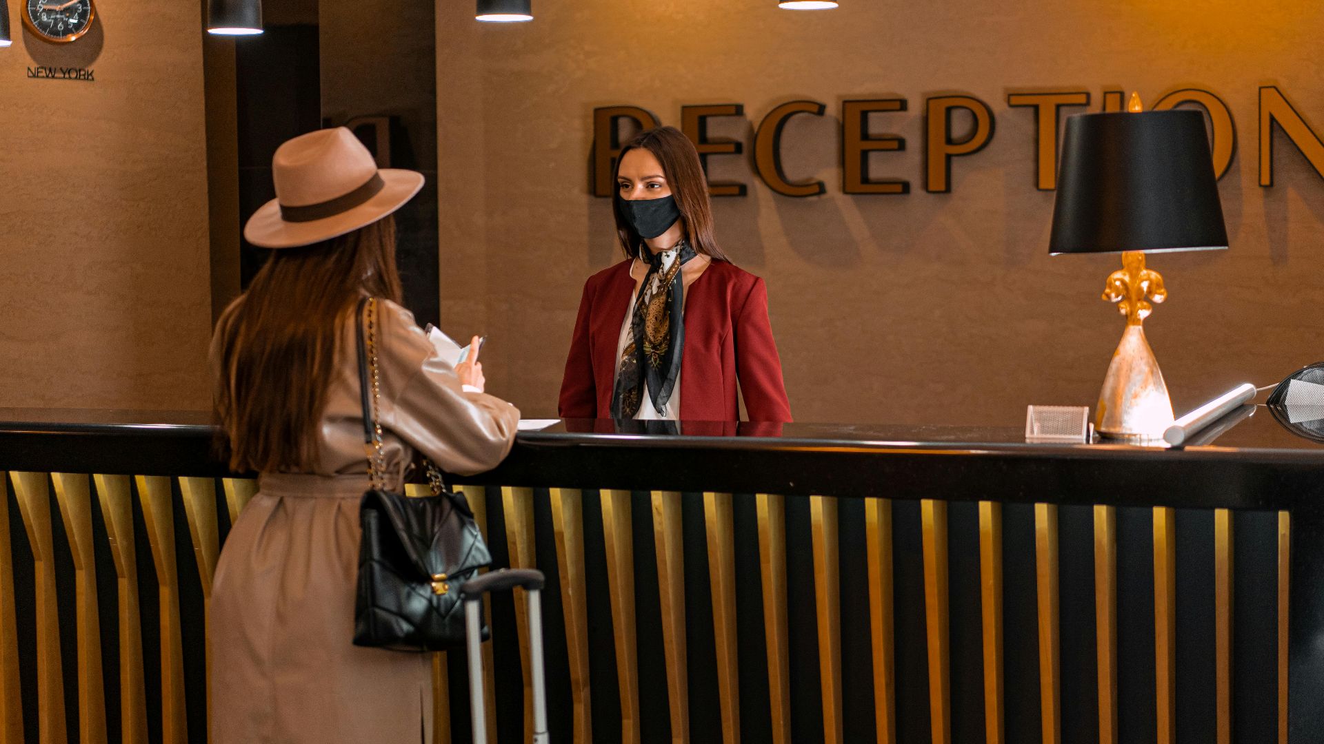 A woman speaks to a receptionist wearing a mask at a hotel reception.