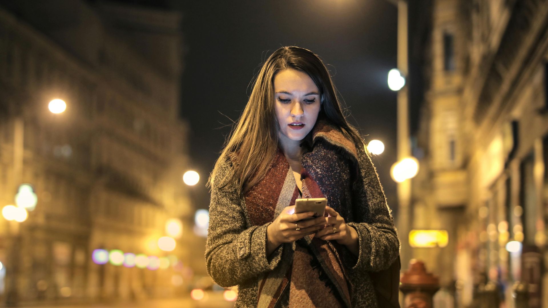 Elegant woman walks through urban street at night, engaged with her smartphone, surrounded by city lights.
