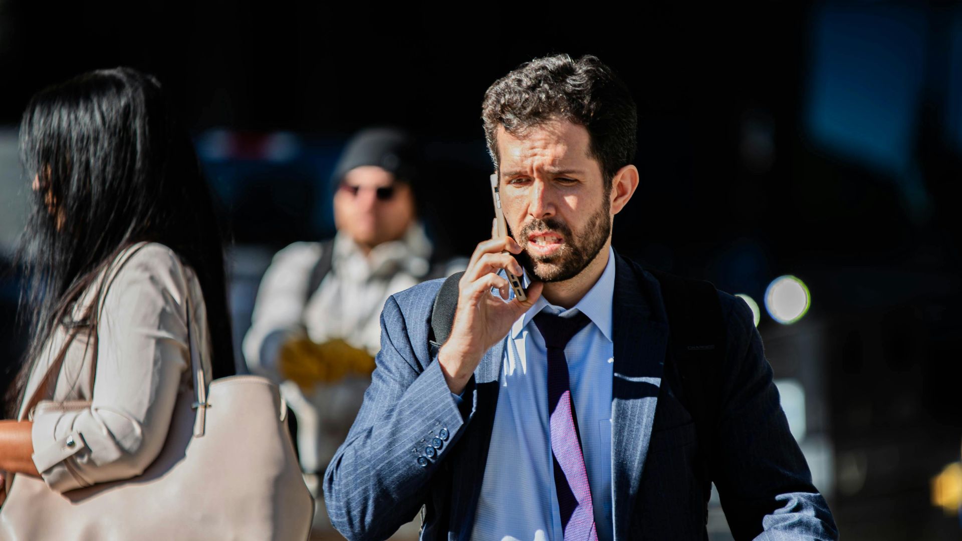 A businessman on a phone call walking with luggage on a sunny city street.