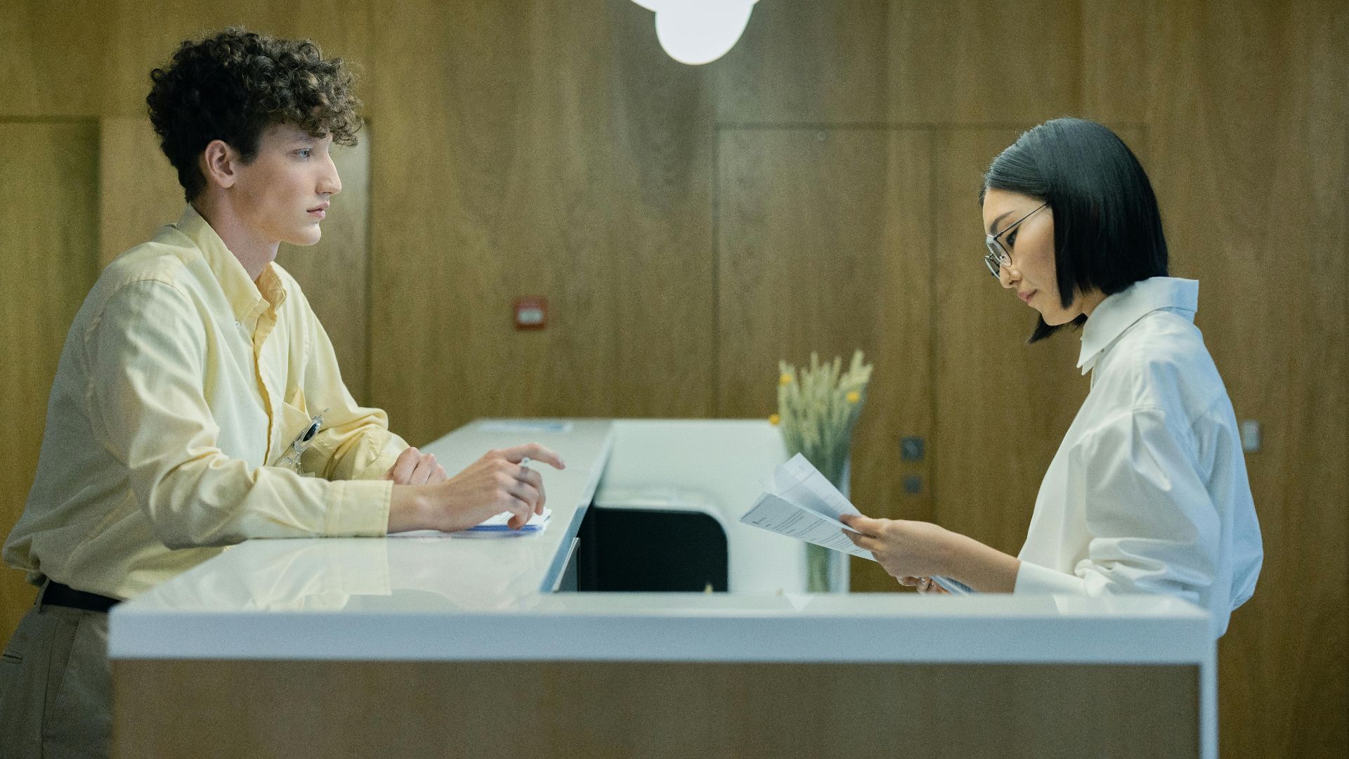 A young man and woman interacting at a modern office reception desk indoors.