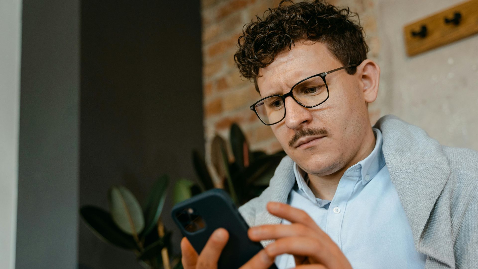 A focused man with glasses using a smartphone indoors next to a brick wall.