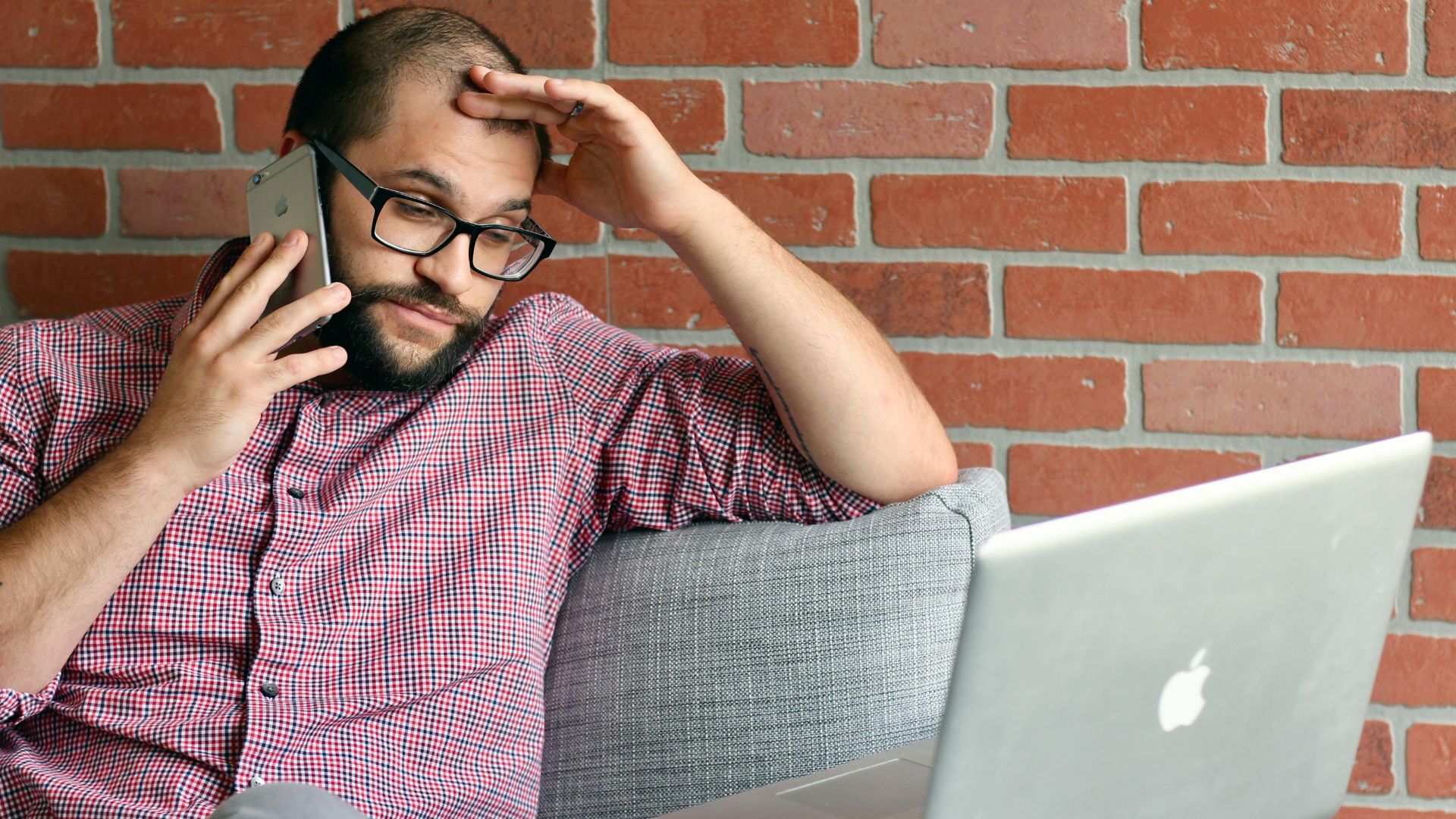 Bearded man looks frustrated while working remotely with laptop and phone.