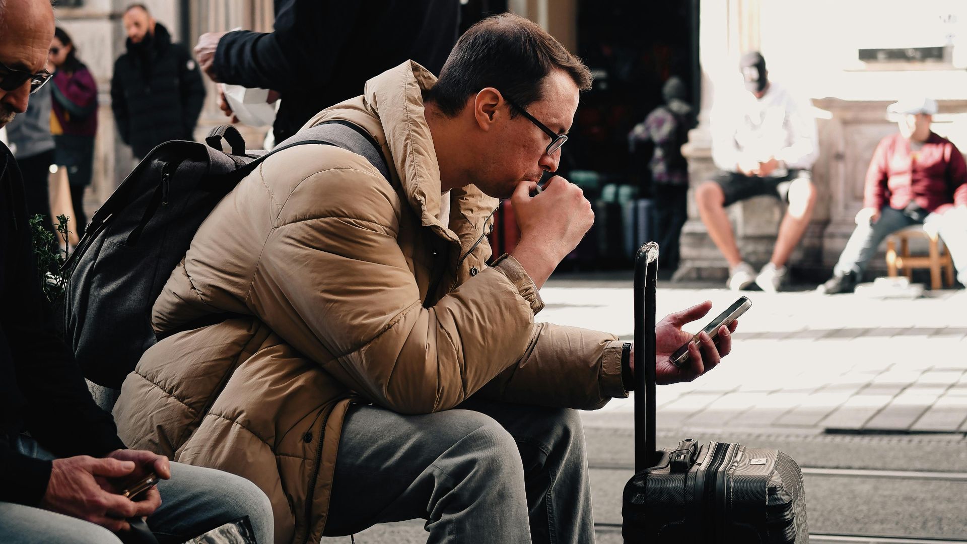A man sitting with luggage, checking his phone in a bustling urban street scene, surrounded by people.