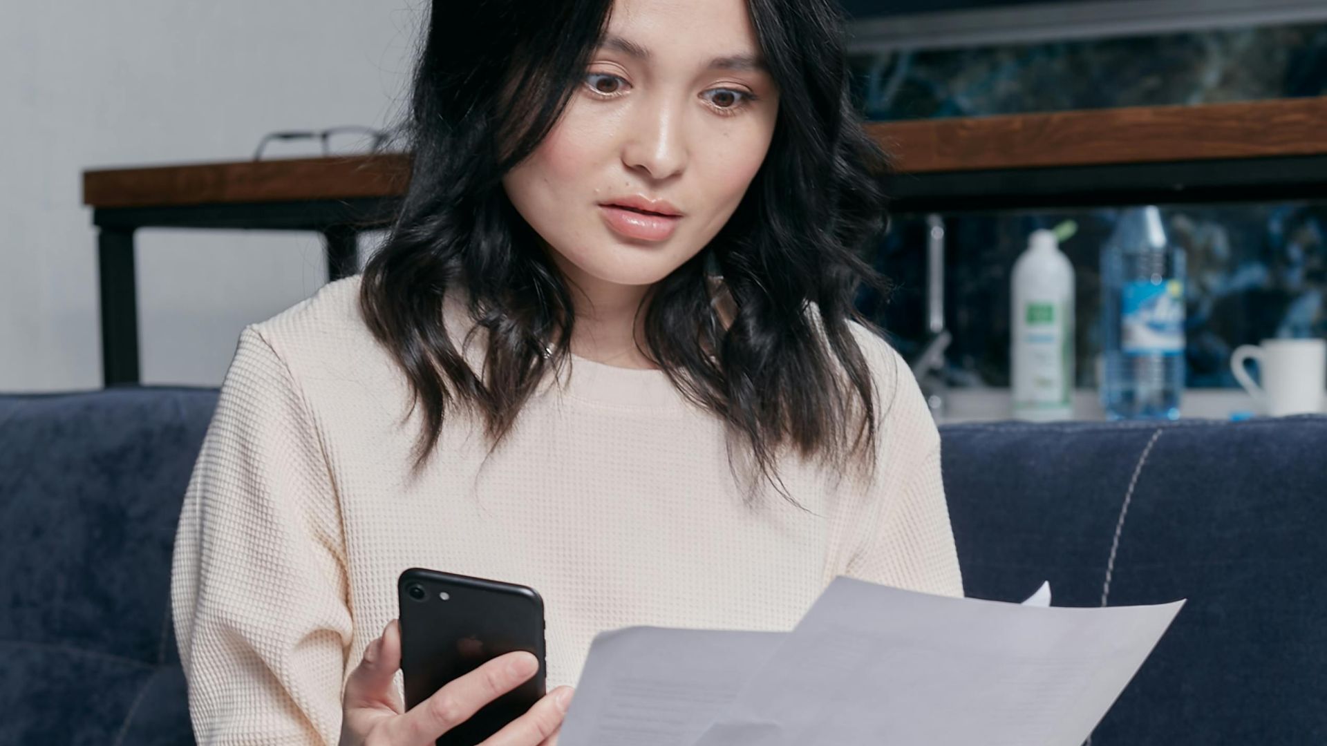 Young woman reviews documents and phone indoors, showcasing concern while seated on a couch with scattered papers. Business concept.