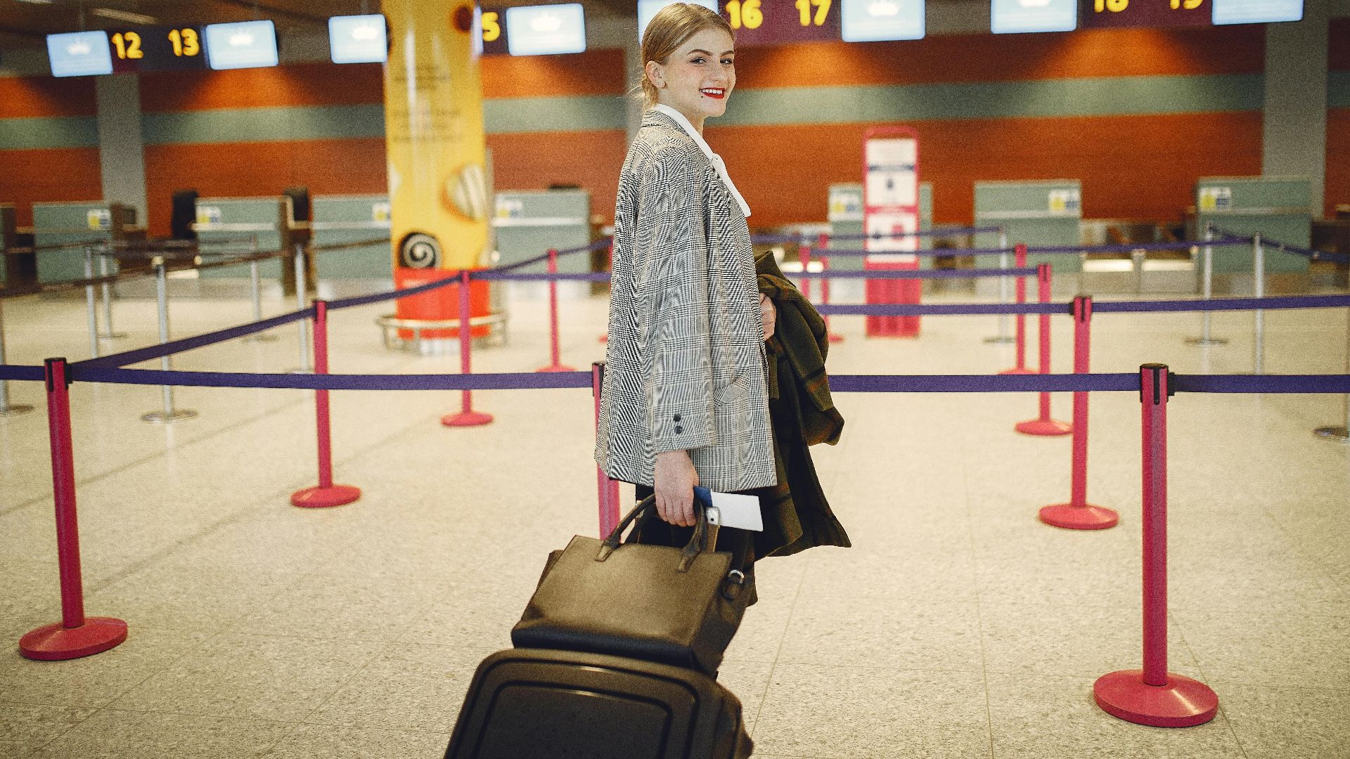 Side view of happy businesswoman in trendy jacket carrying baggage and smiling at camera while waiting for flight in modern airport