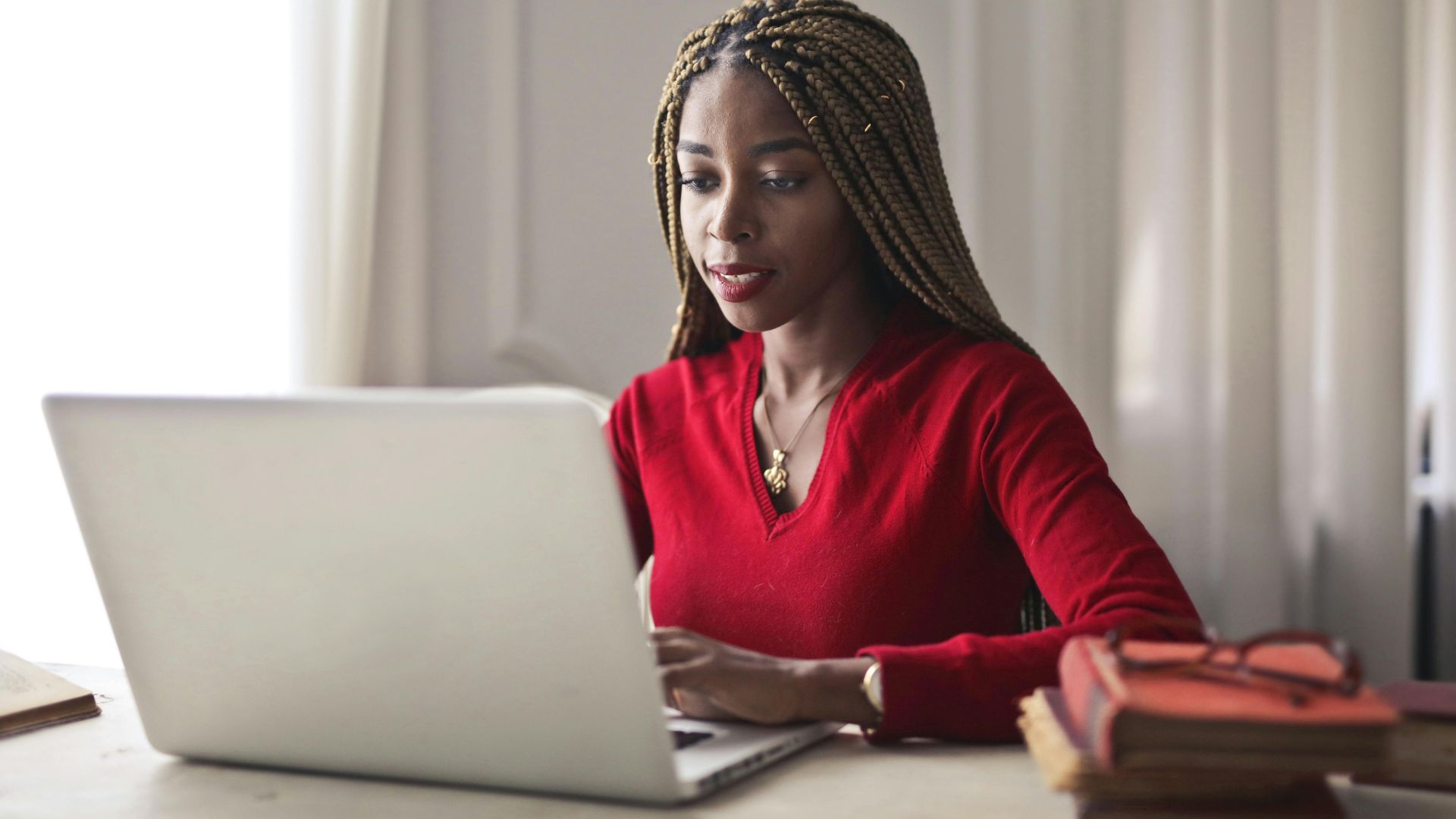 Confident woman in a red sweater working on a laptop in a bright, organized workspace.