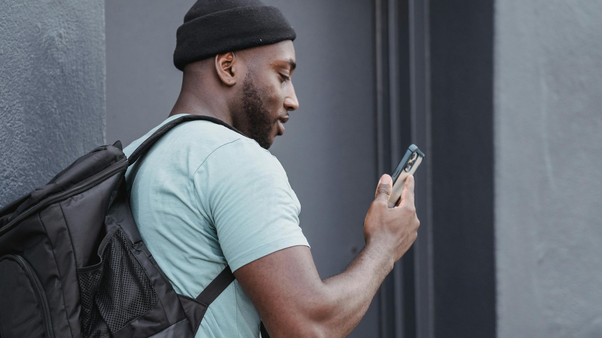 Courier with a backpack and scooter checks phone beside a building outdoors.
