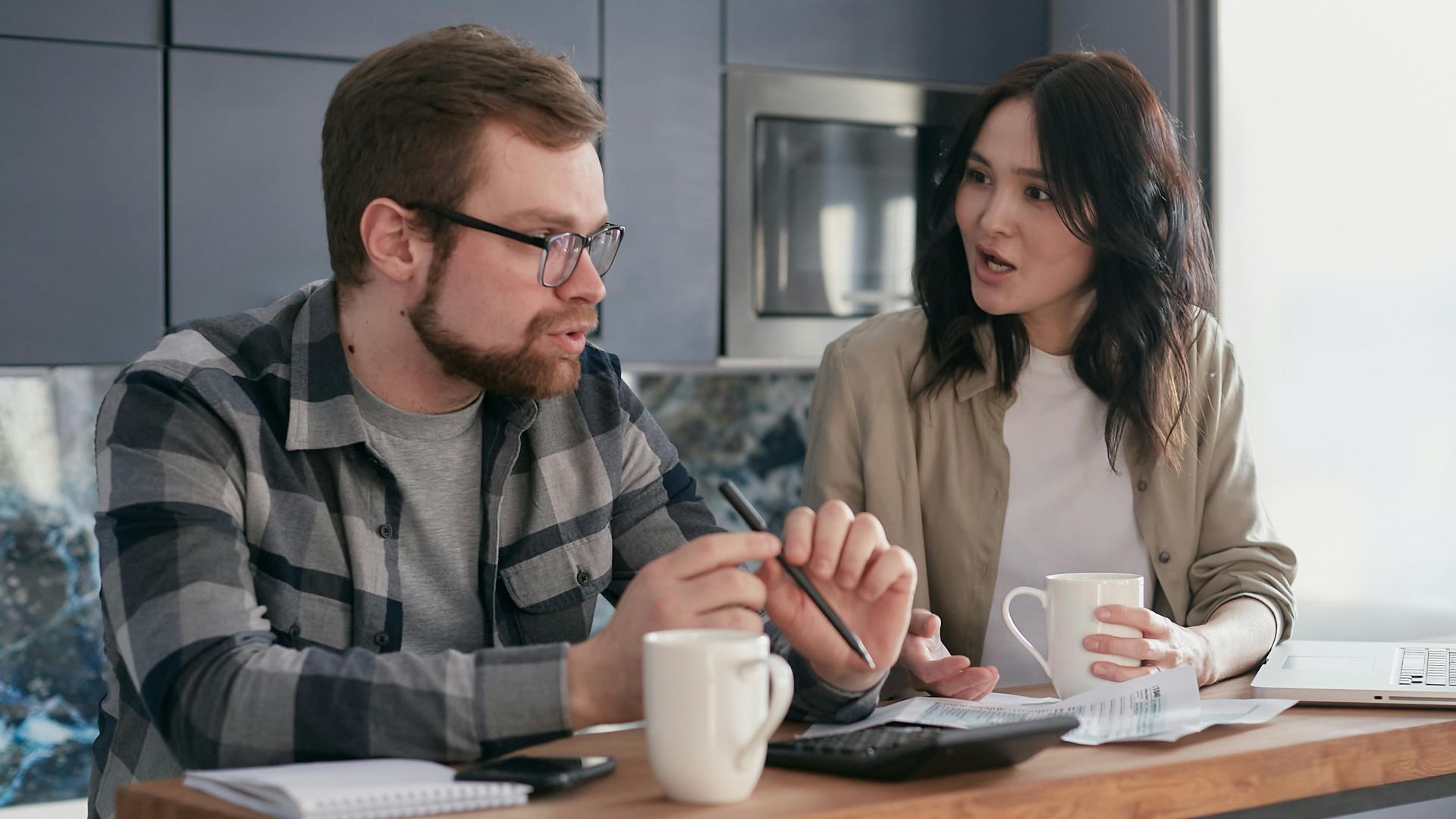 A couple managing finances with gadgets, documents, and coffee mugs at home.