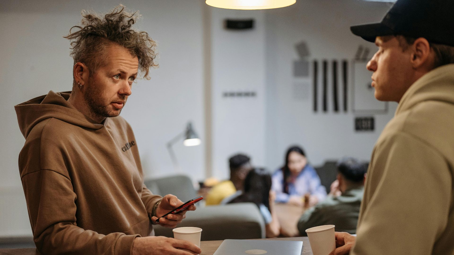 Two men in a casual office setting talking over coffee with laptops.