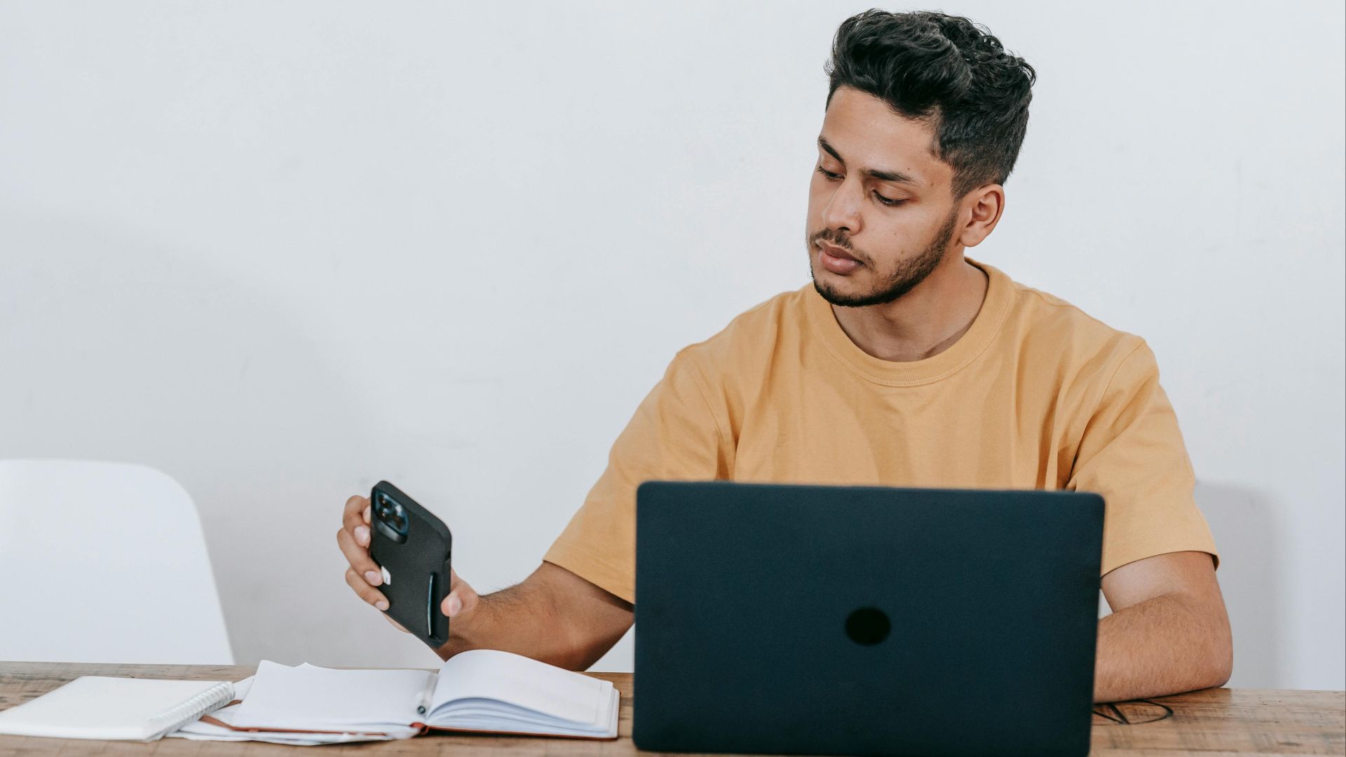 Serious young bearded Hispanic male entrepreneur in casual clothes checking notifications on mobile phone while working at table with laptop and notebook at workplace