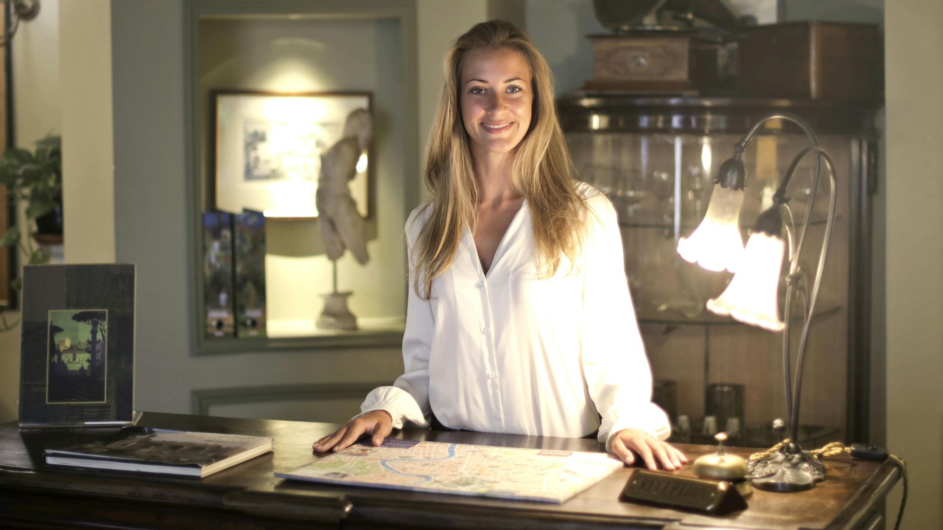 Smiling female receptionist wearing elegant clothes standing at reception desk in old fashioned interior of hotel and looking at camera