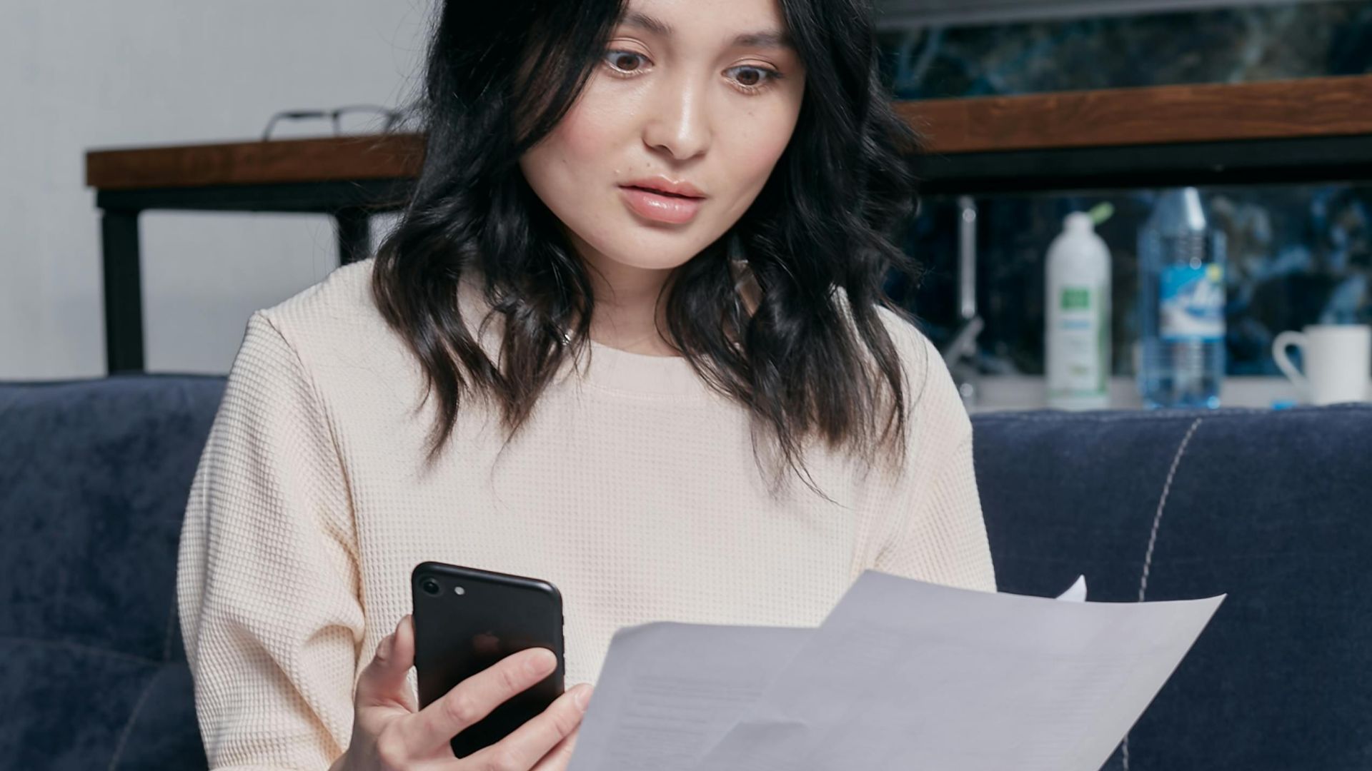 Young woman reviews documents and phone indoors, showcasing concern while seated on a couch with scattered papers. Business concept.