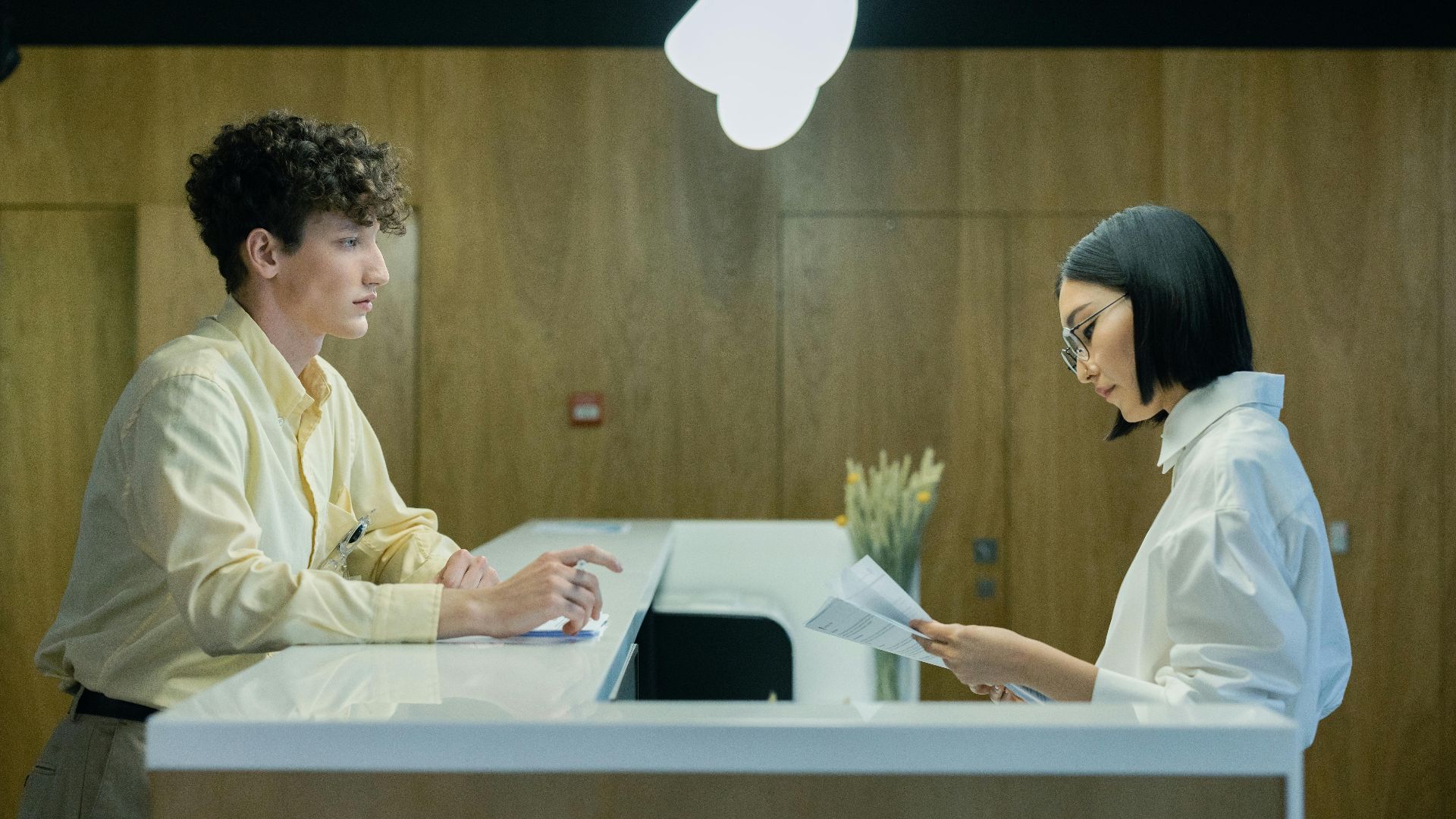 A young man and woman interacting at a modern office reception desk indoors.
