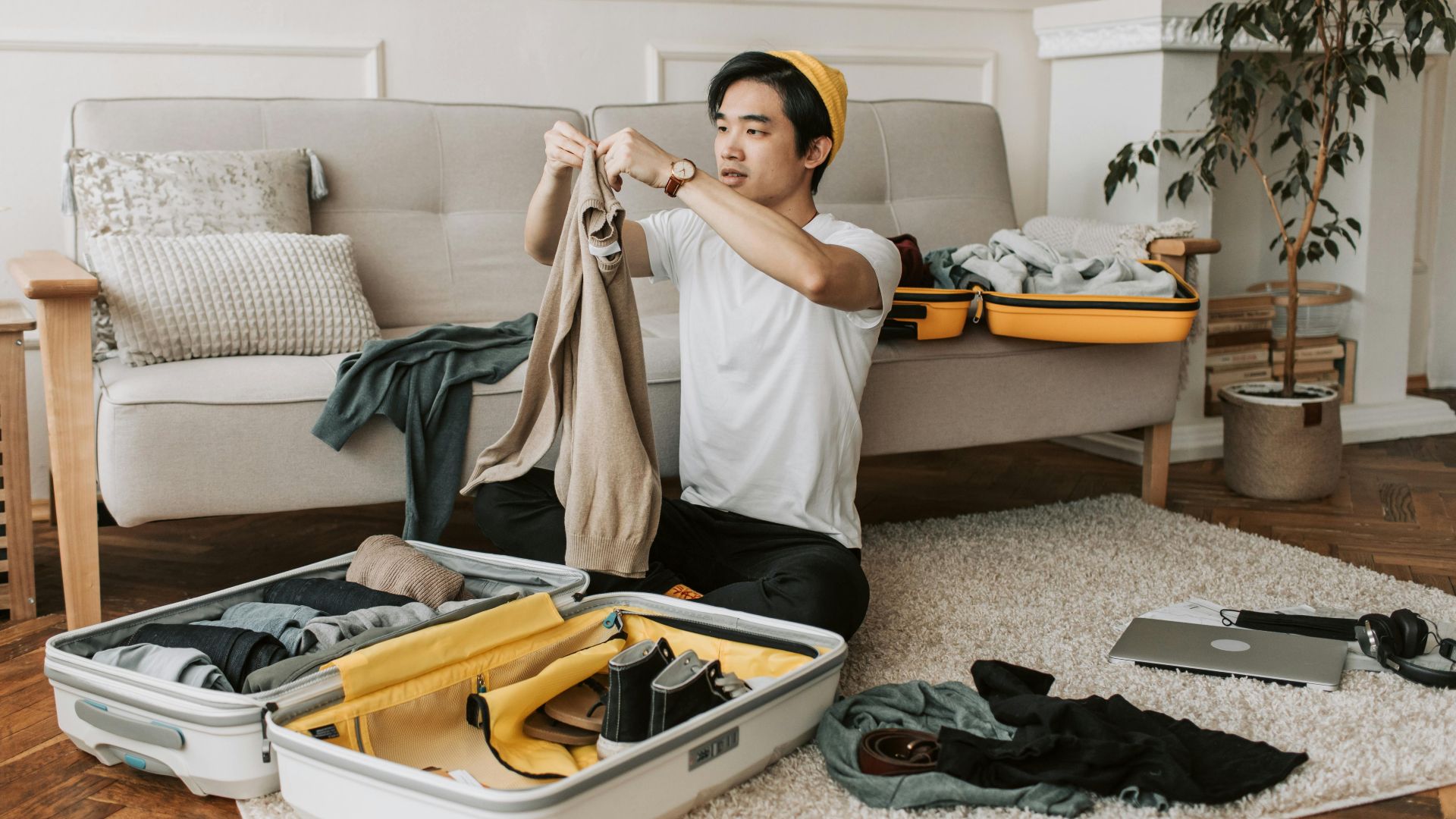 A young man organizes clothes into a suitcase in a stylish living room, preparing for a trip.