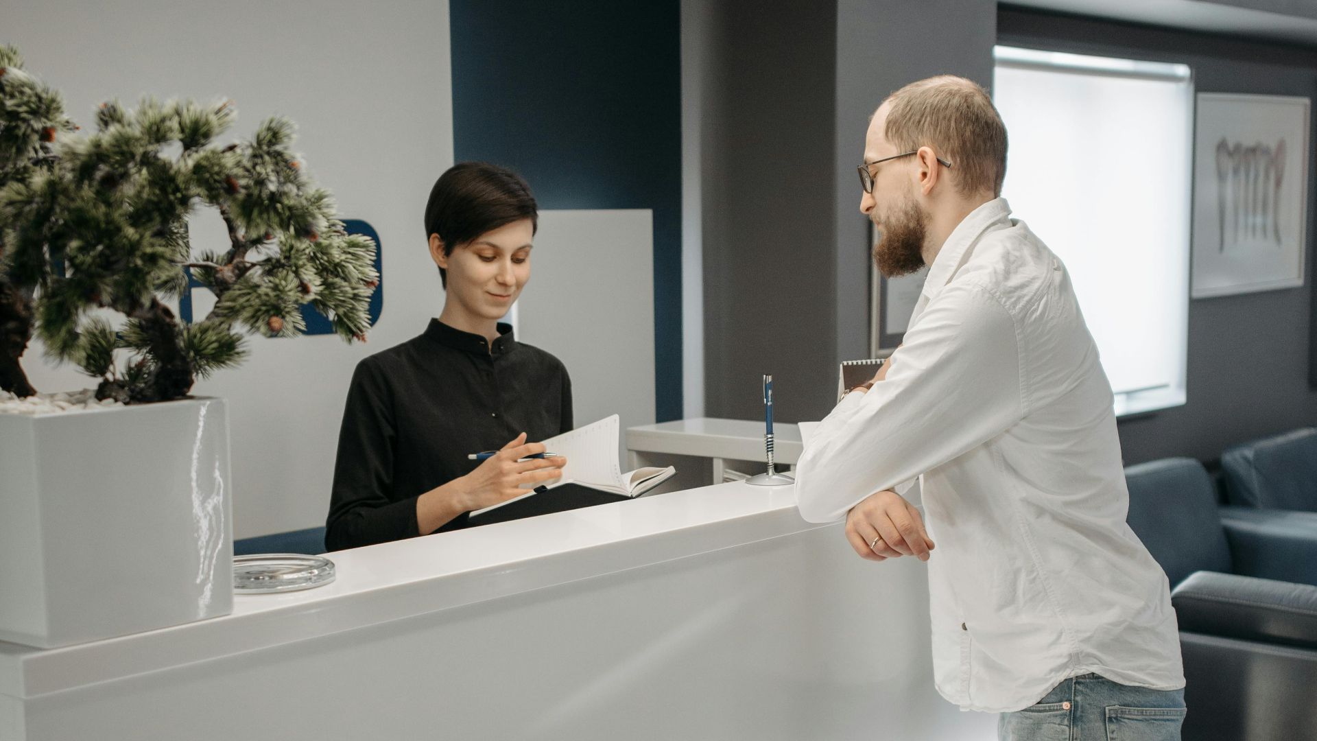 A man and woman at a sleek office reception desk, discussing paperwork in a professional setting.