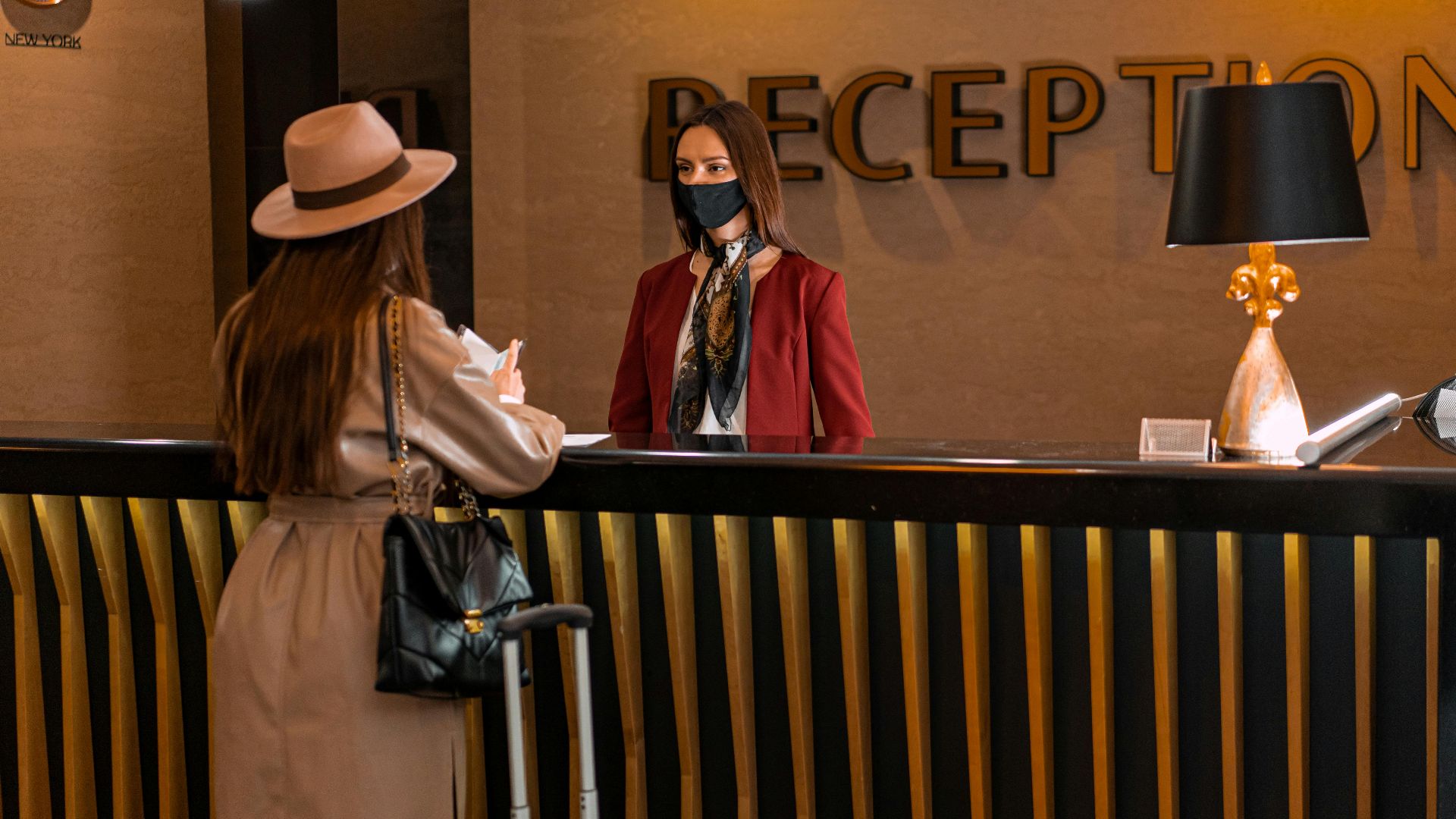 A woman speaks to a receptionist wearing a mask at a hotel reception.