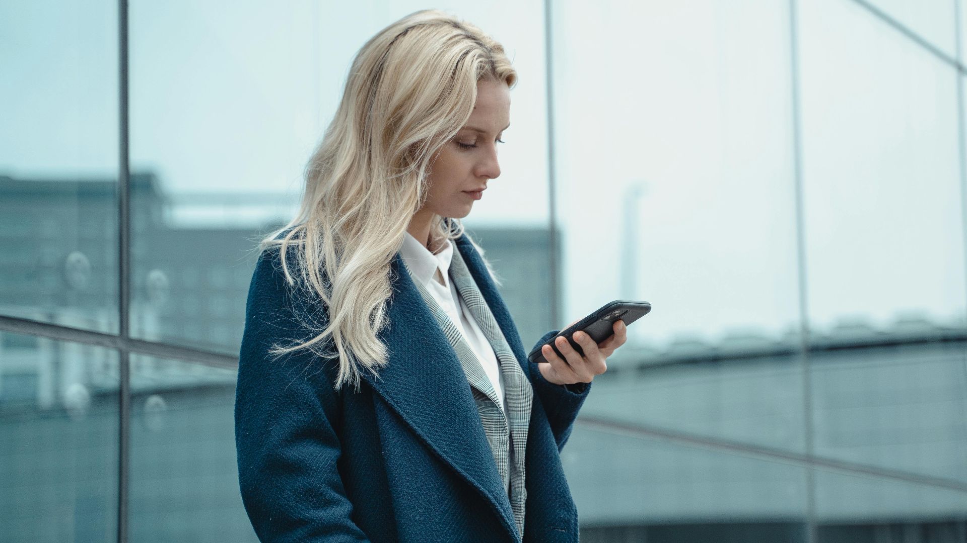Professional woman in blue coat using smartphone outside office building.