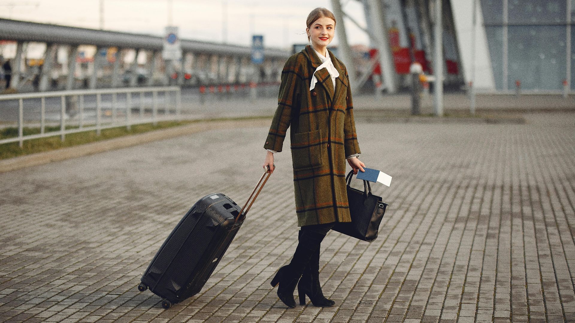 Full body young female passenger in trendy plaid coat with suitcase strolling on pavement near modern building of ariport terminal taking bag and documents