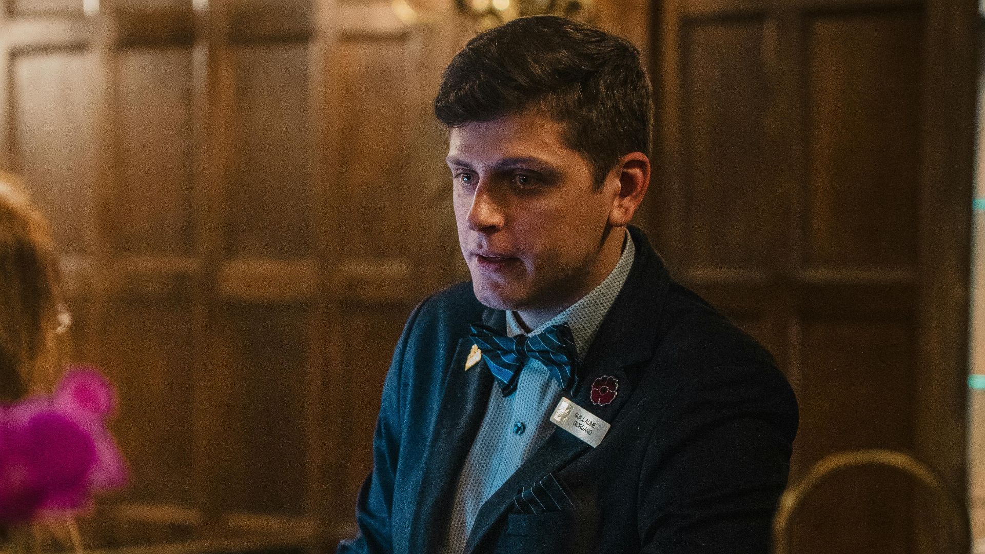 Polite hotel receptionist in formal attire at a warmly lit wooden reception desk.