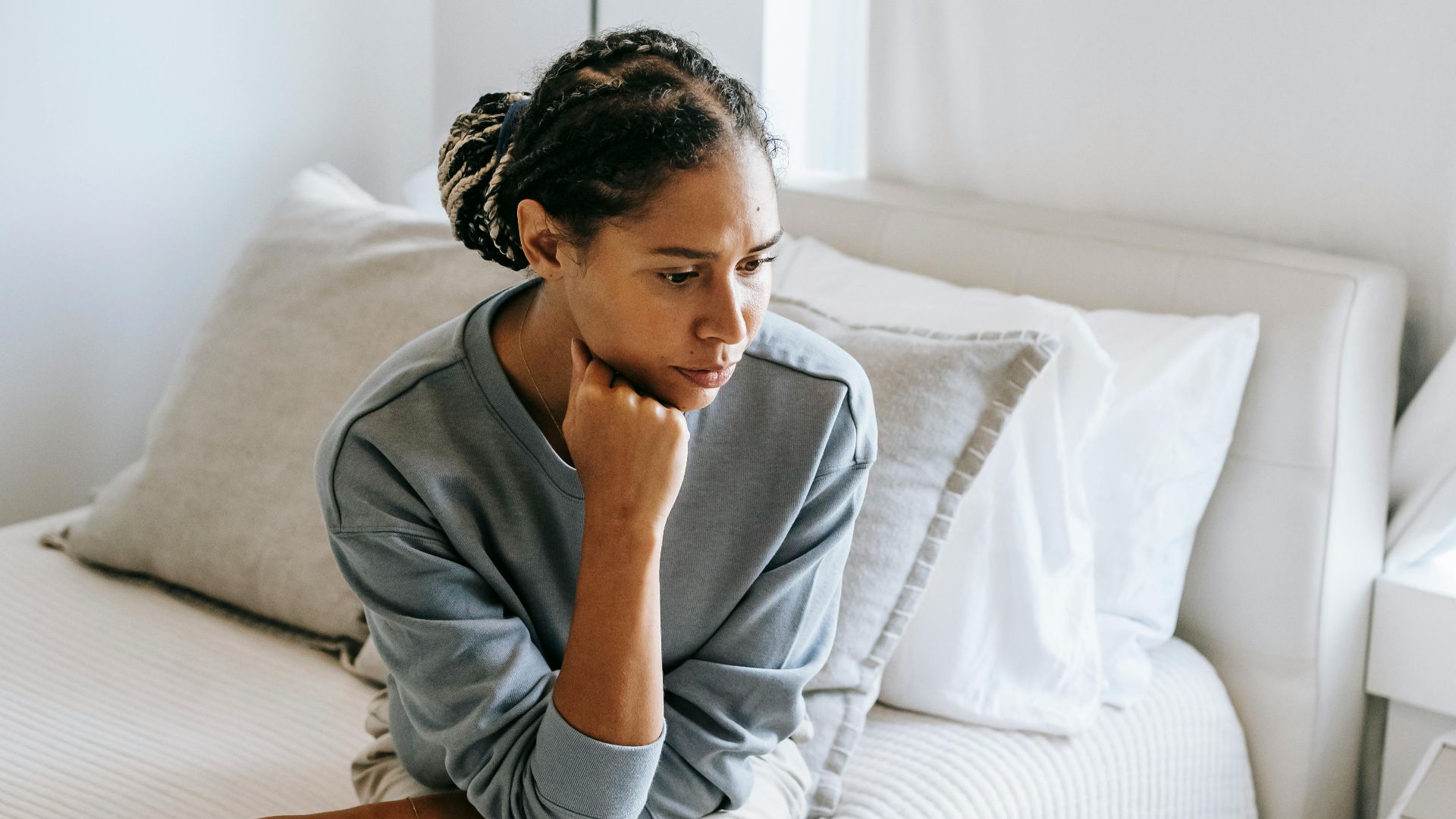 Serious young African American lady in casual clothes sitting on white bed and thinking while looking down in bedroom
