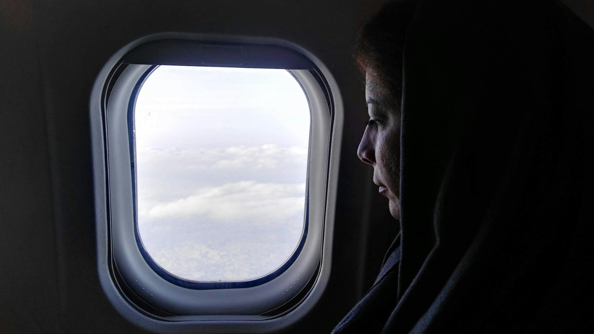 A woman in a hijab looks pensively out an airplane window at the clouds below.