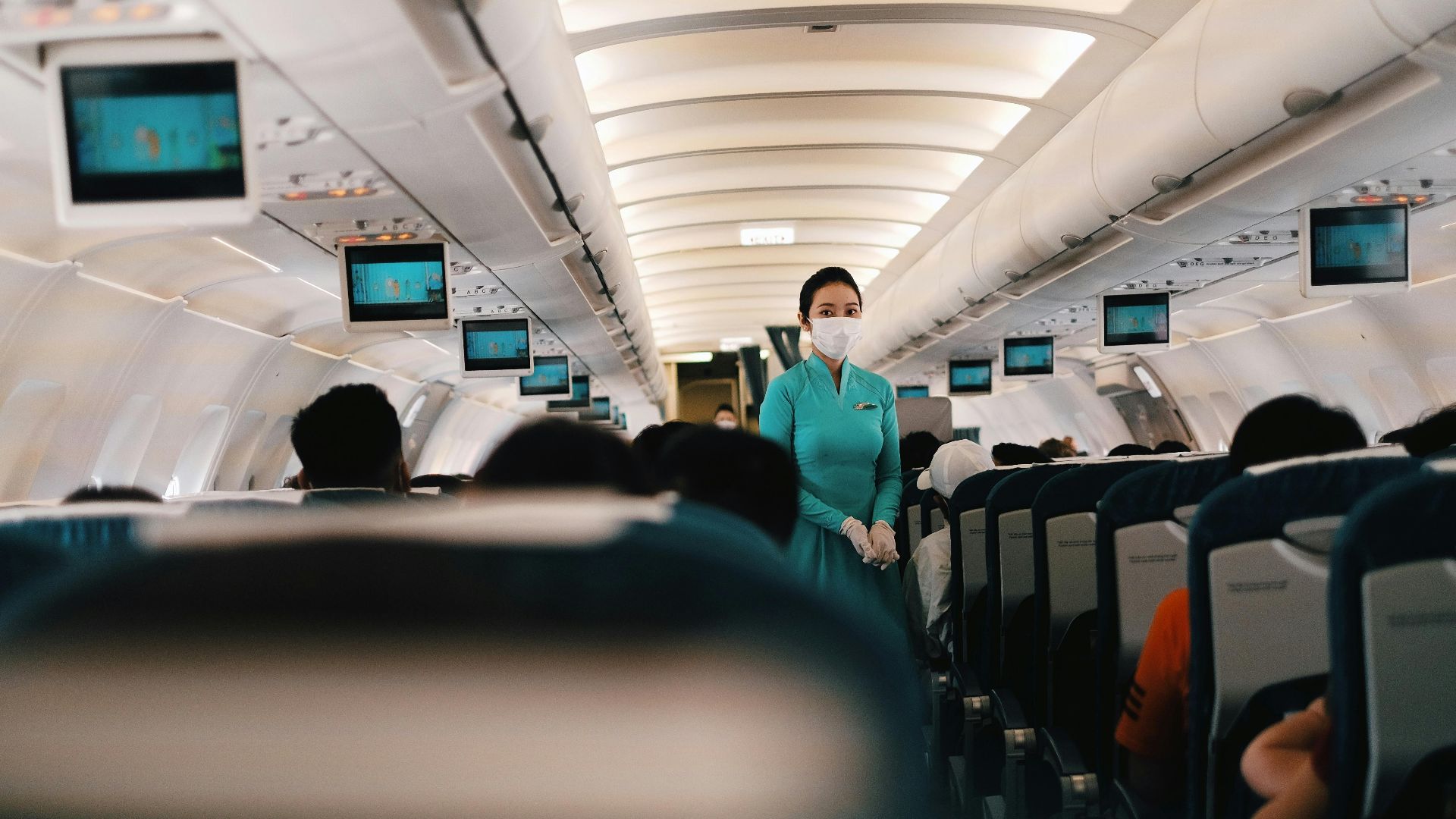Flight attendant wearing a face mask attending to passengers in an airplane cabin.