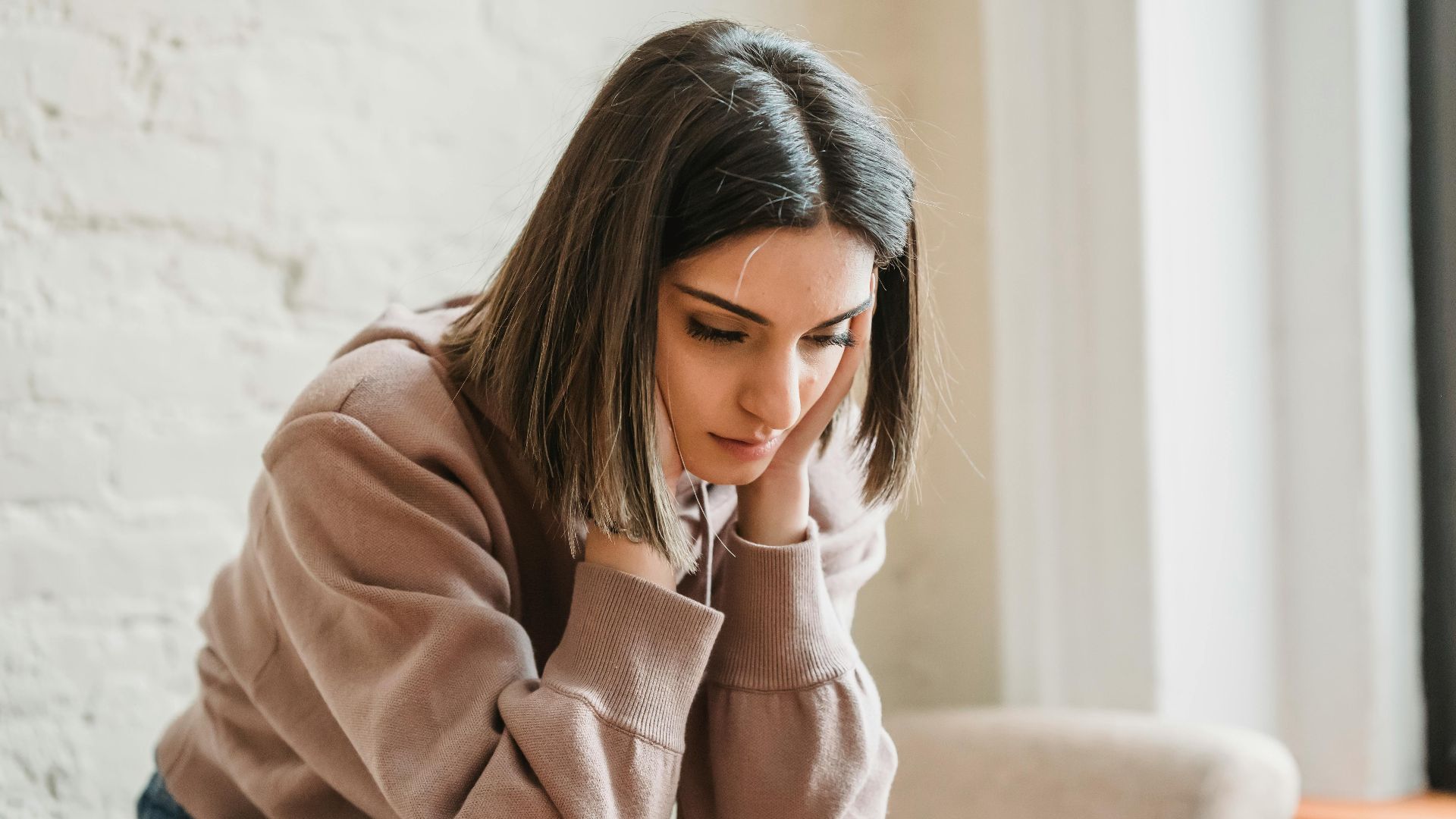 Unhappy female in casual wear touching face and looking down while sitting on sofa in light living room at home