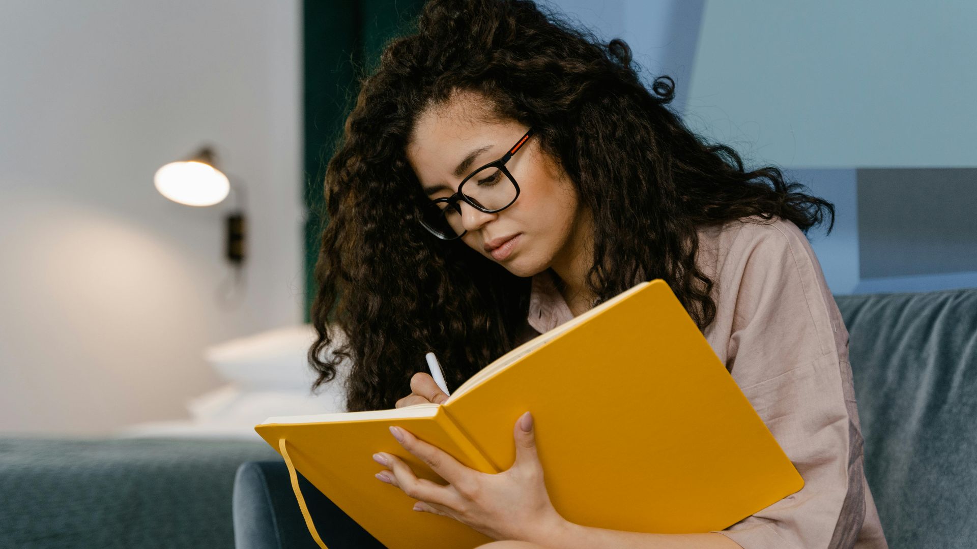 Young woman with curly hair writes in a yellow notebook, seated indoors.