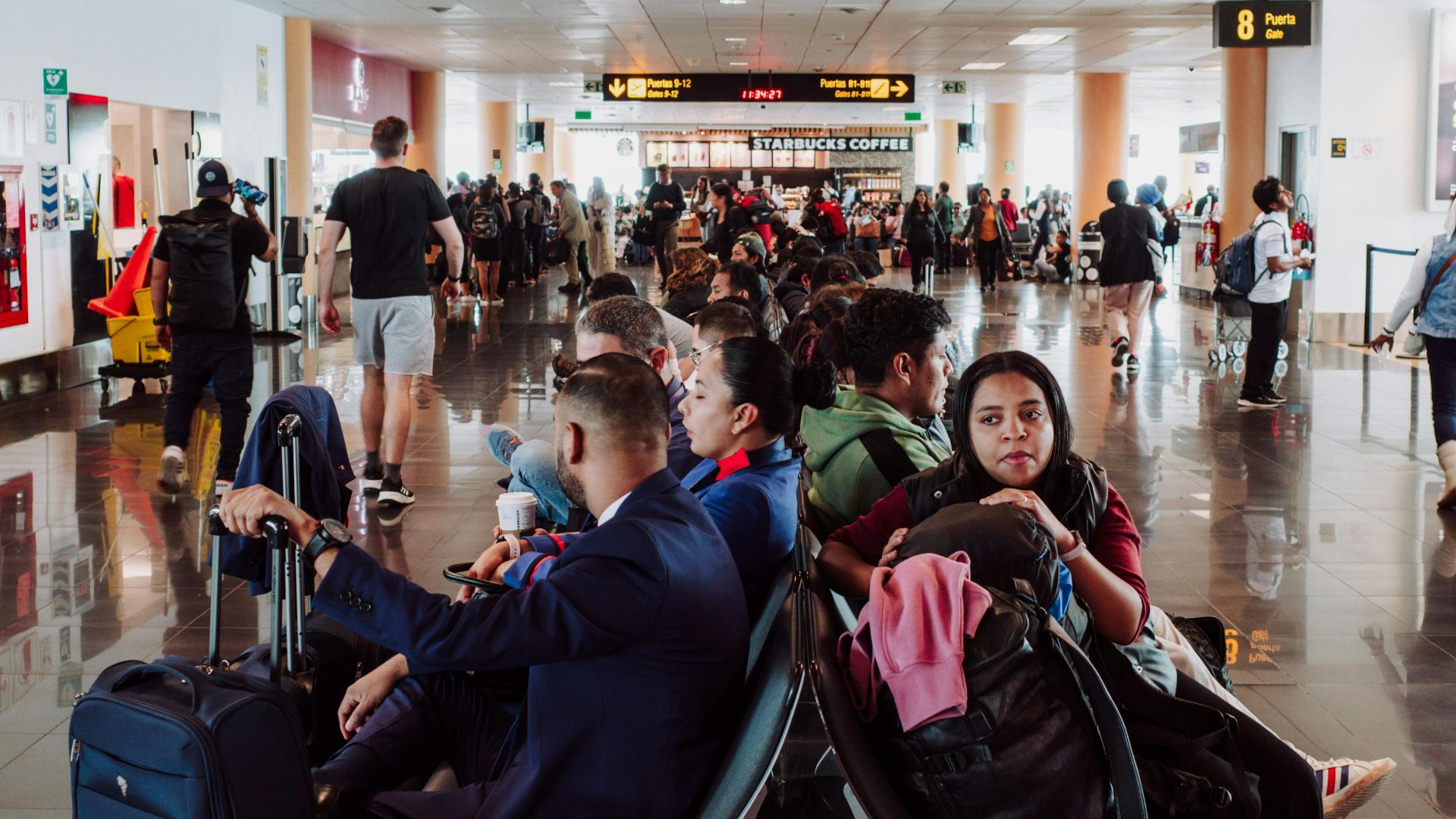 Passengers waiting in a busy airport terminal seating area, with luggage in the modern airport.