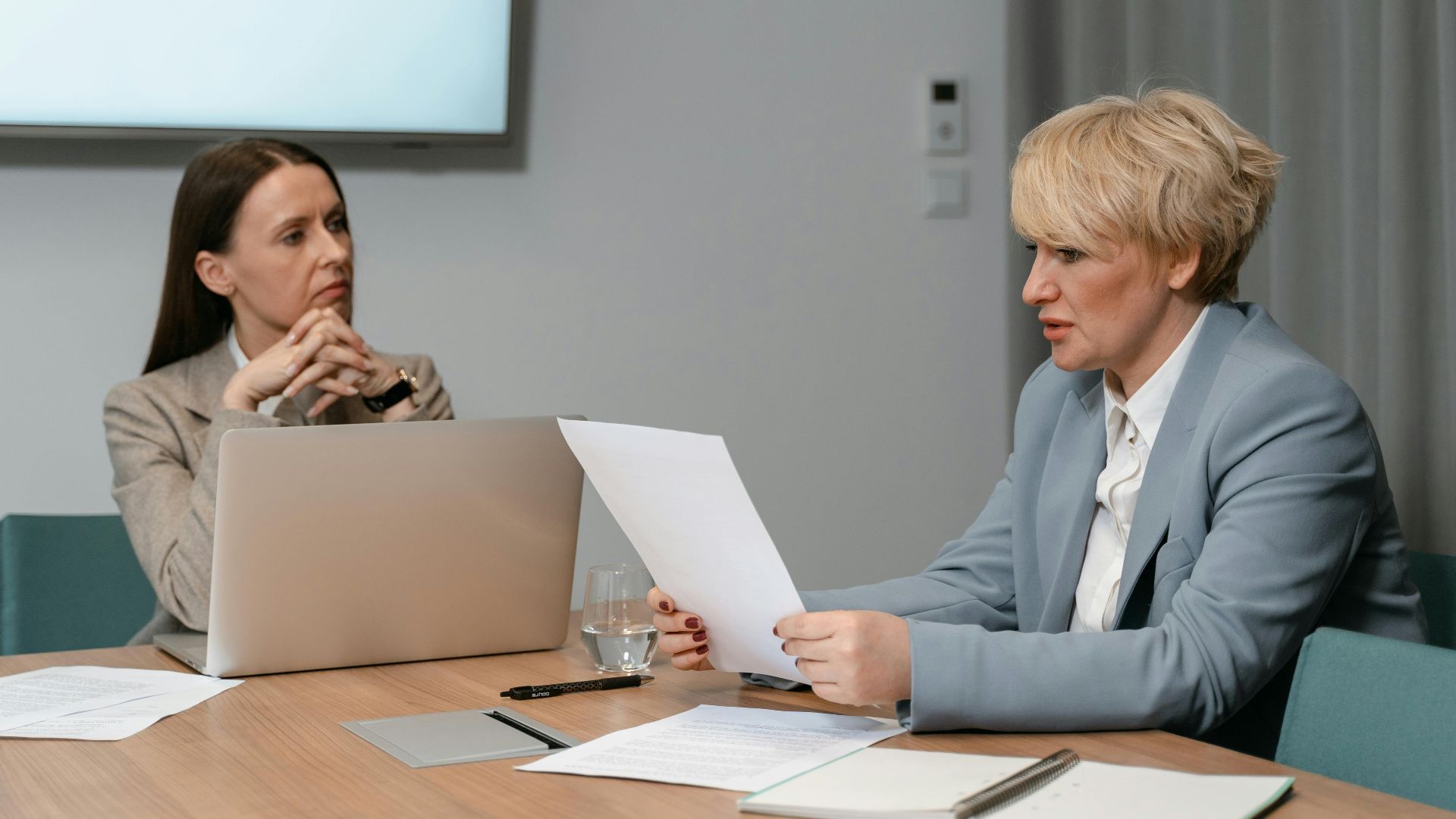 Two businesswomen in a serious meeting, discussing documents with a laptop in an office setting.