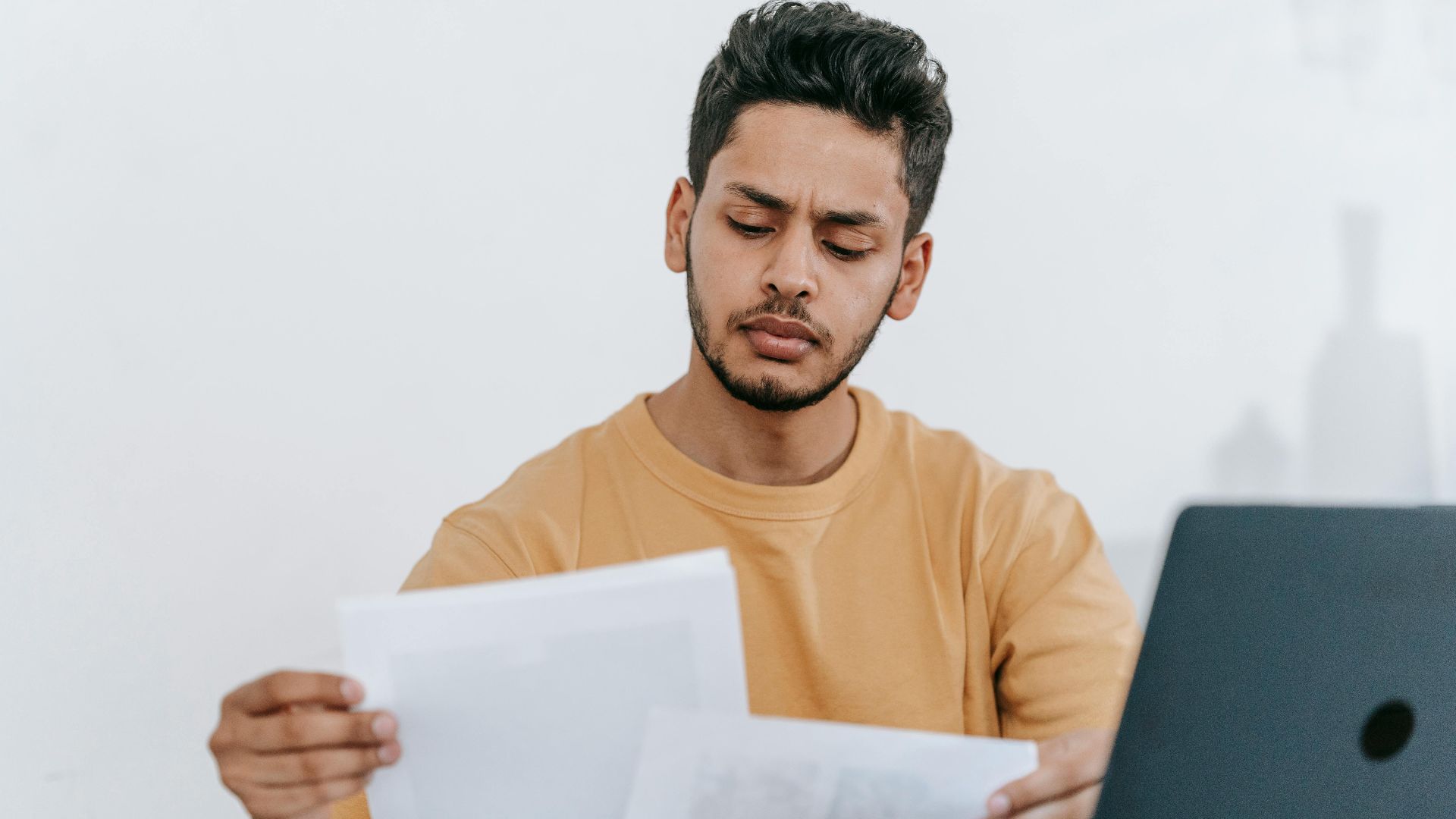 Attentive young bearded Hispanic male remote worker examining papers with results of project while working with laptop in home office