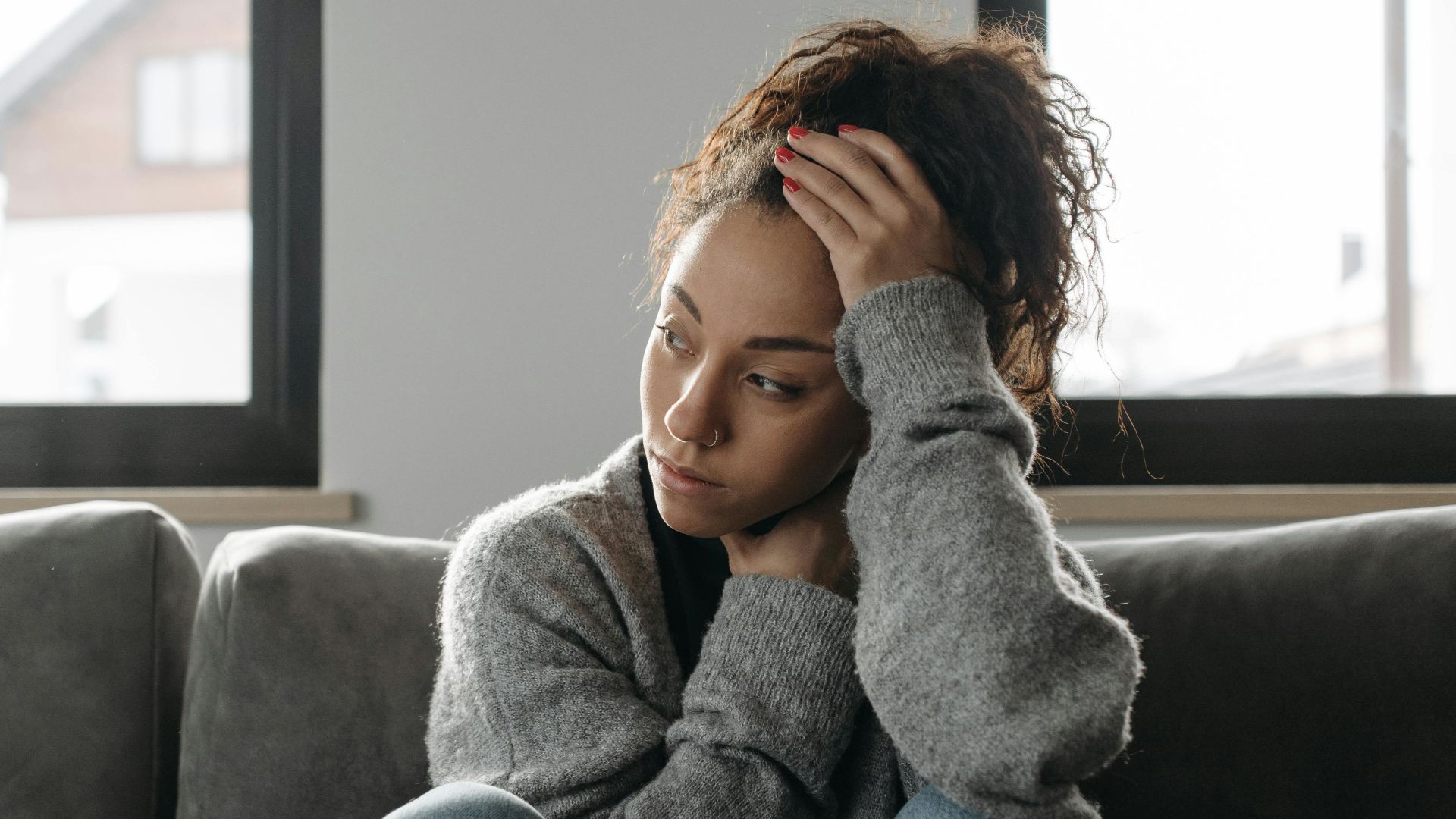 Thoughtful woman in cozy sweater sitting on sofa indoors by the window.