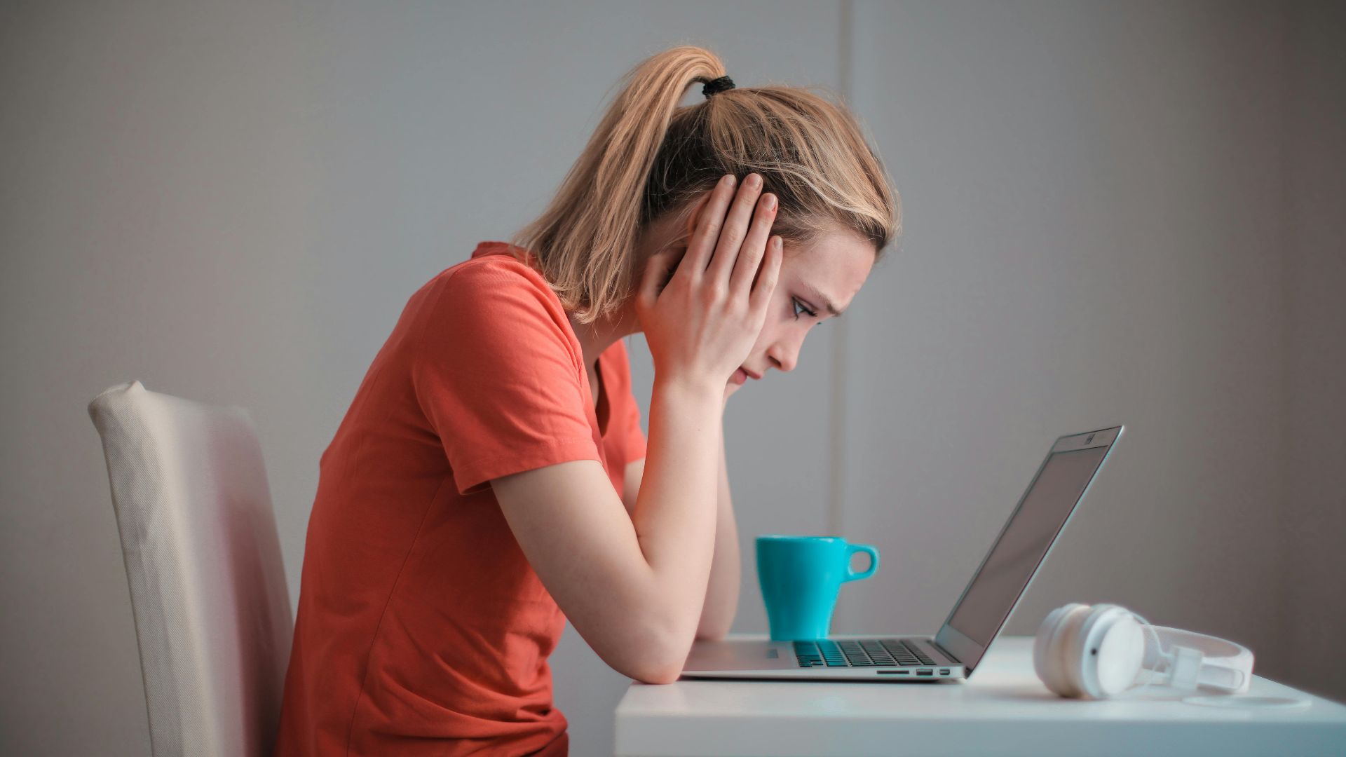 Side view of thoughtful worried female student in casual clothes focusing on screen and trying to solve problem while sitting at table with blue mug of hot drink and headphones and working on laptop at home