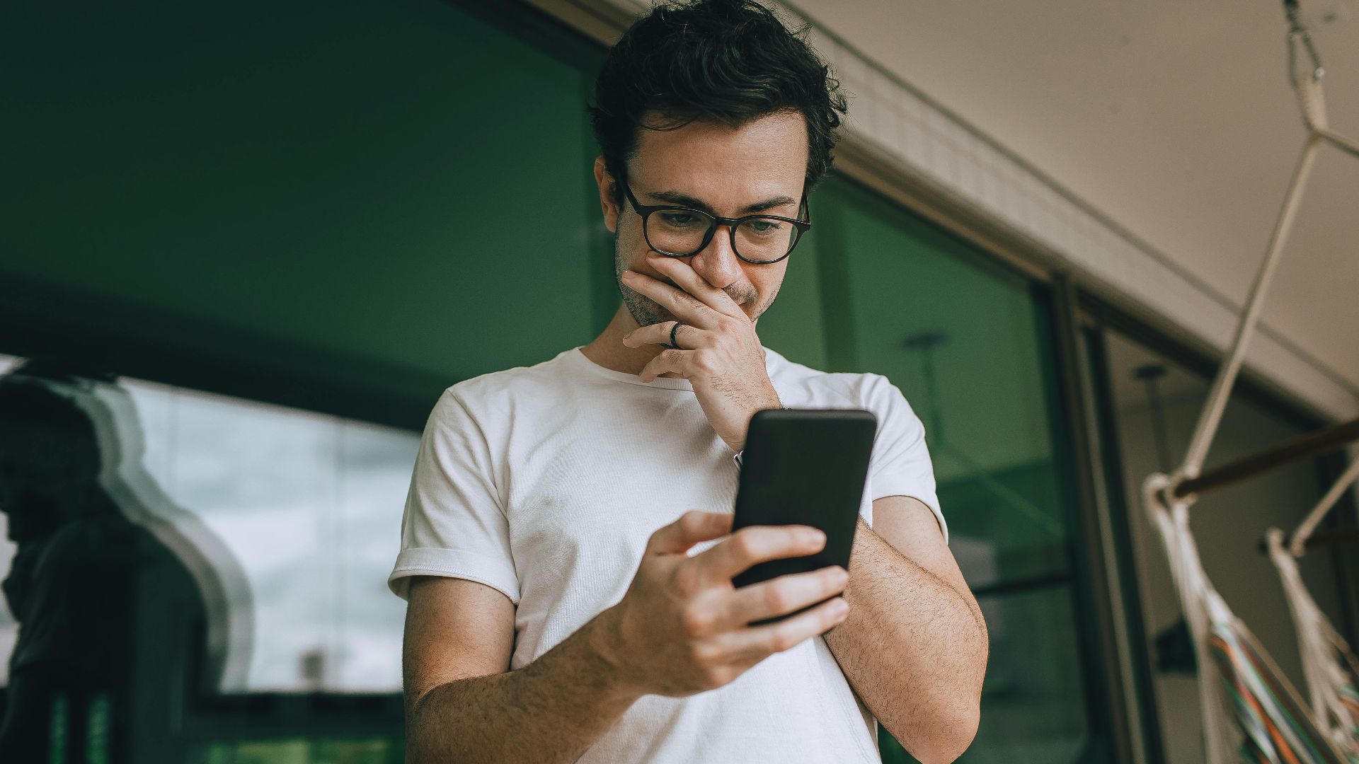 A young man in glasses standing on a balcony, using his smartphone with a thoughtful expression.