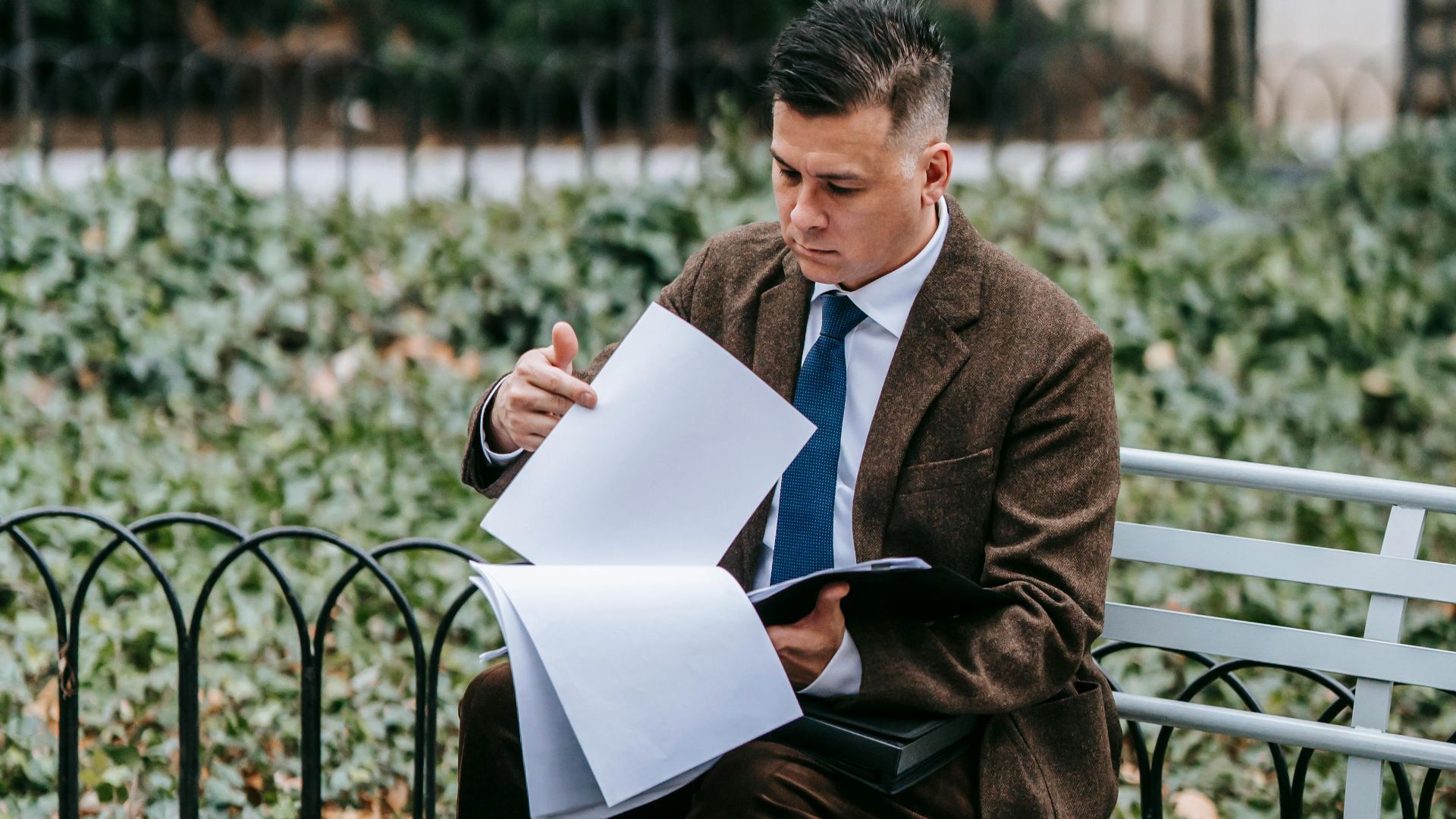 Professional man in formal wear reviewing documents while sitting on a park bench outdoors.
