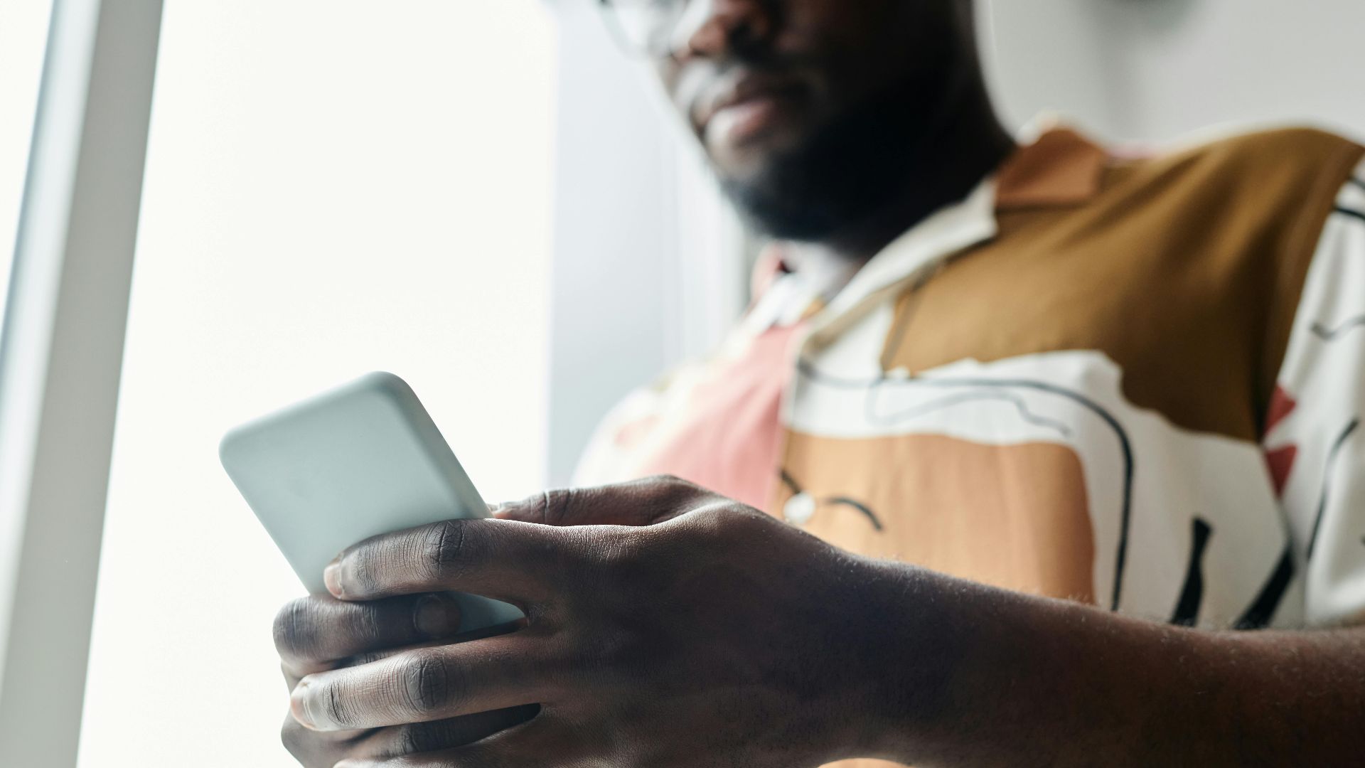 A close-up of a man using a smartphone by a window, featuring a modern patterned shirt.