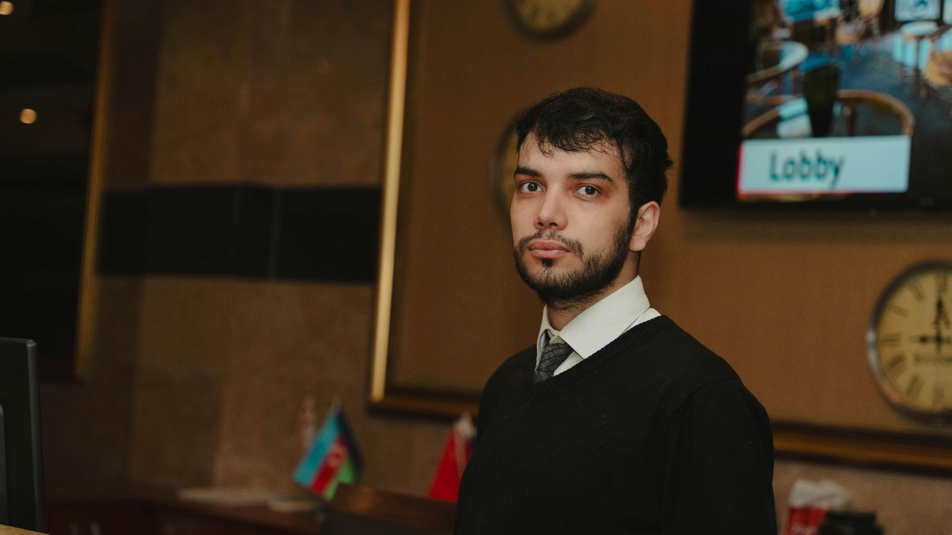 A hotel receptionist standing at the front desk in a hotel lobby, ready to assist guests.