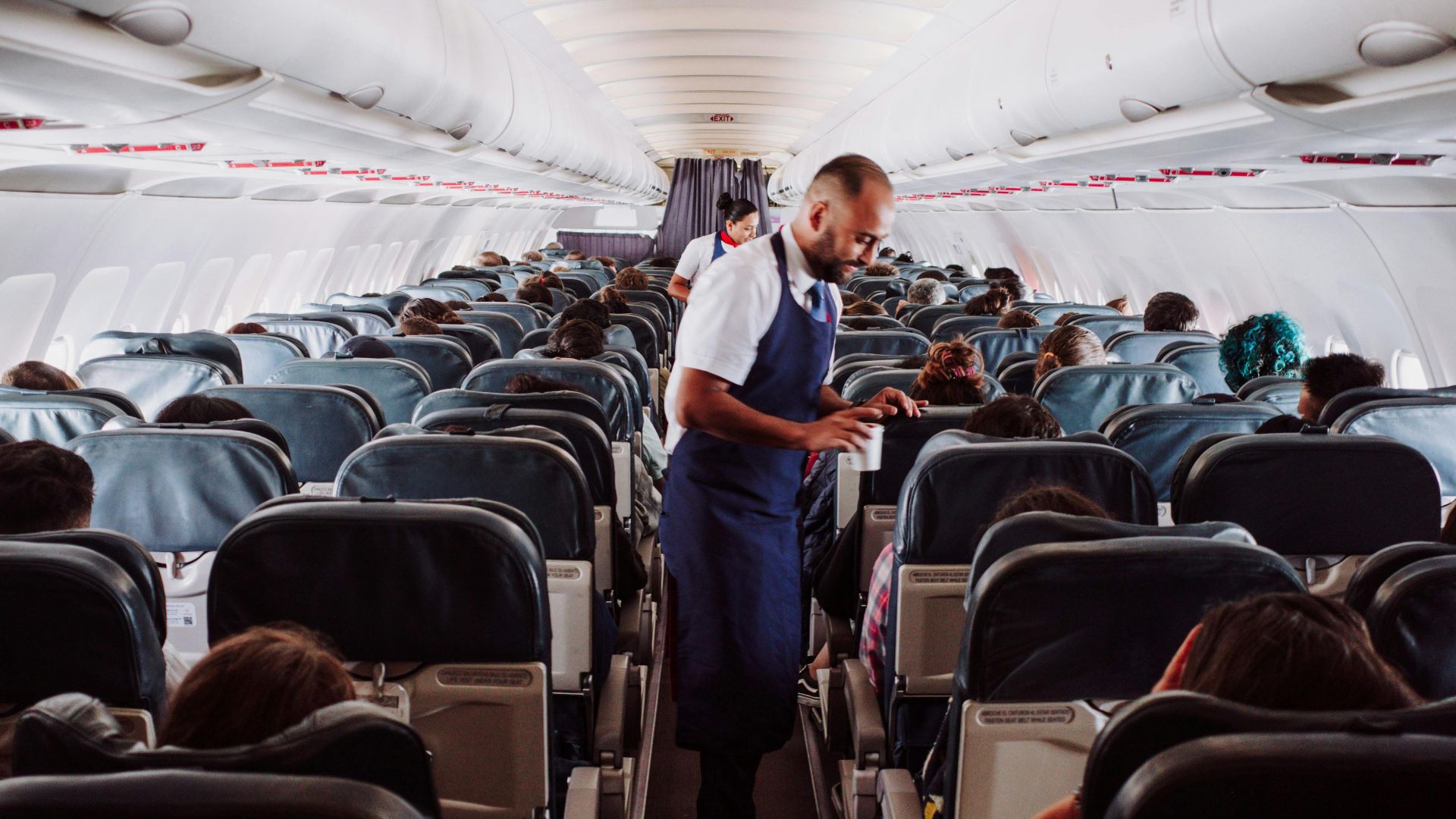 Flight attendant assists passengers in a crowded airplane aisle with rows of seats.
