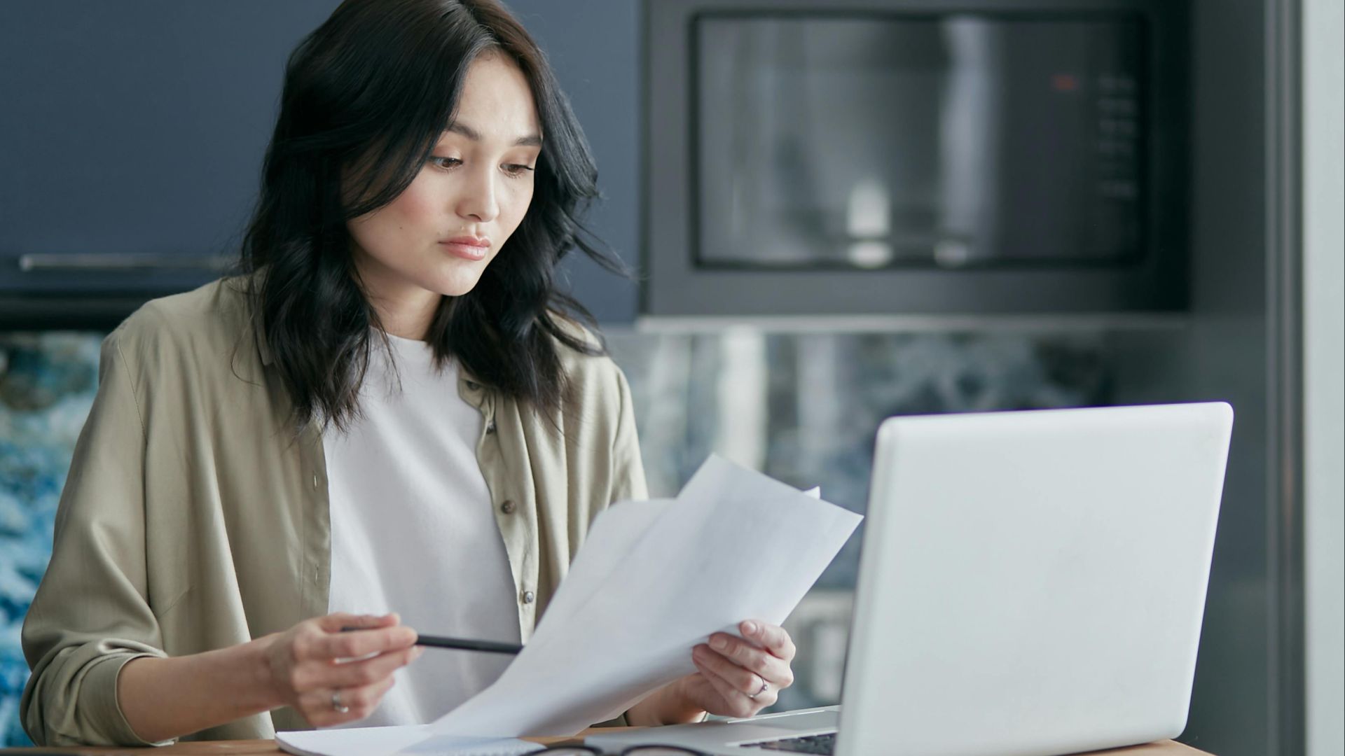 Concentrated woman reviewing documents and working on a laptop at home office setup in kitchen.