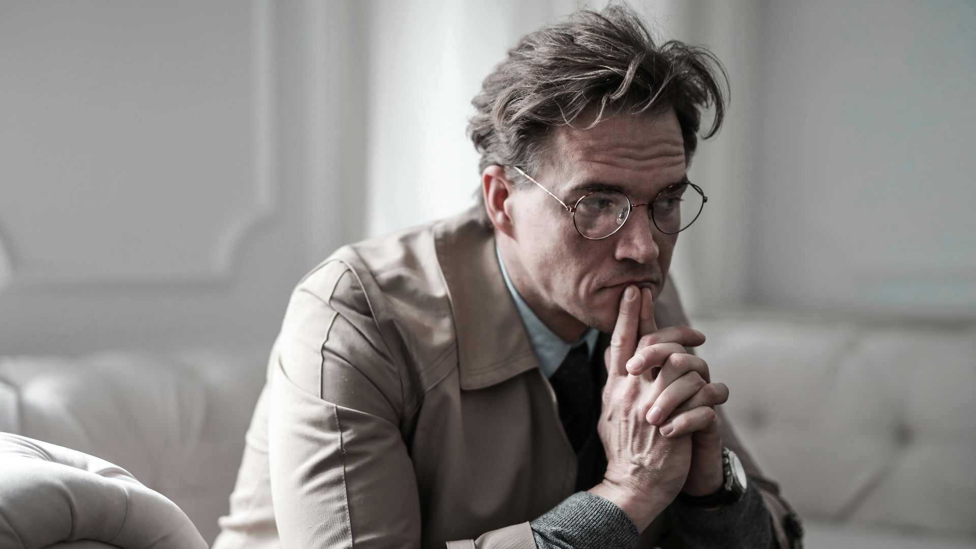 A thoughtful man wearing glasses sits on a sofa in a classic interior, lost in deep contemplation.