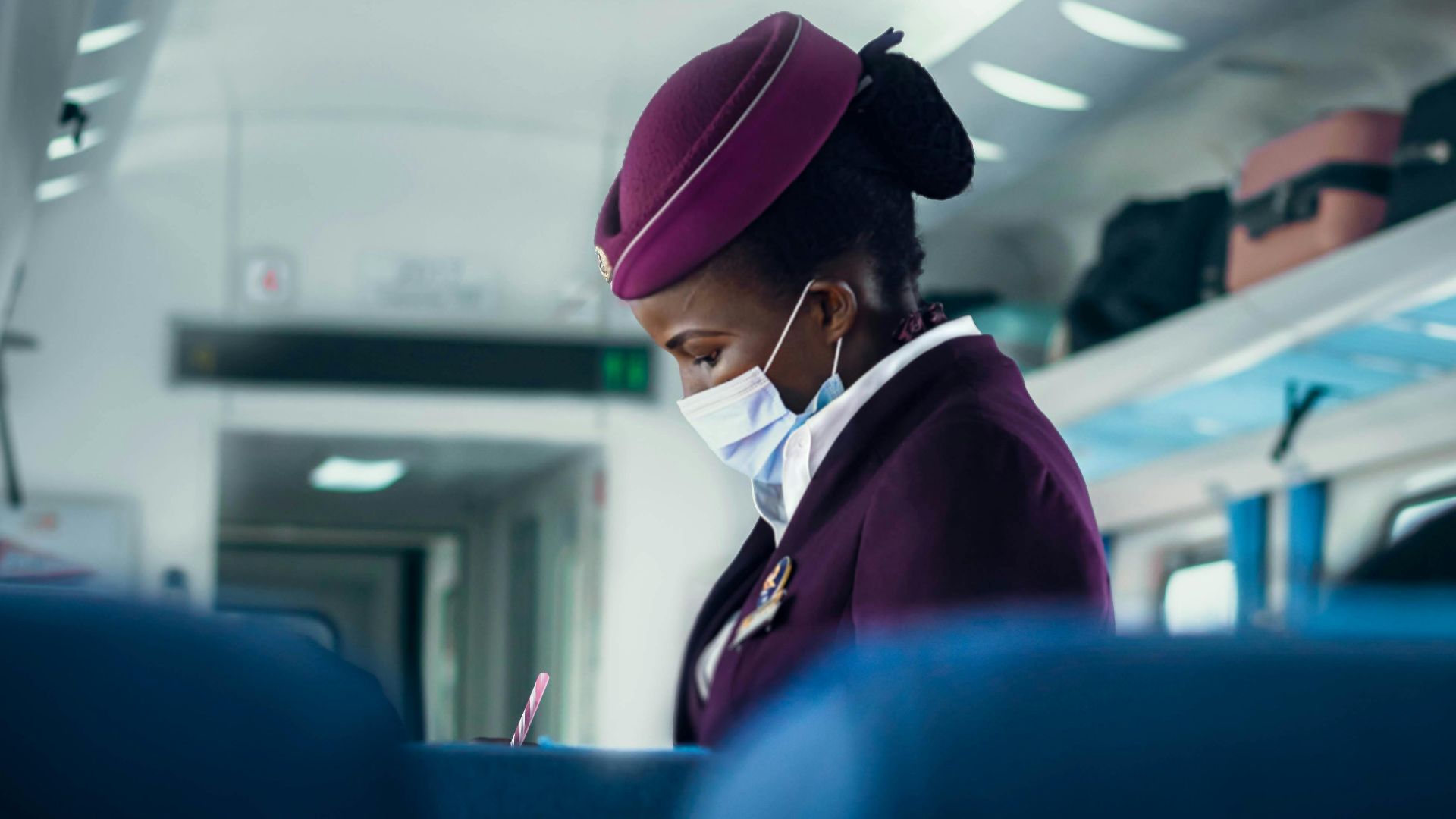 A flight attendant in a purple uniform wearing a surgical mask, attending to passengers in an airplane cabin.