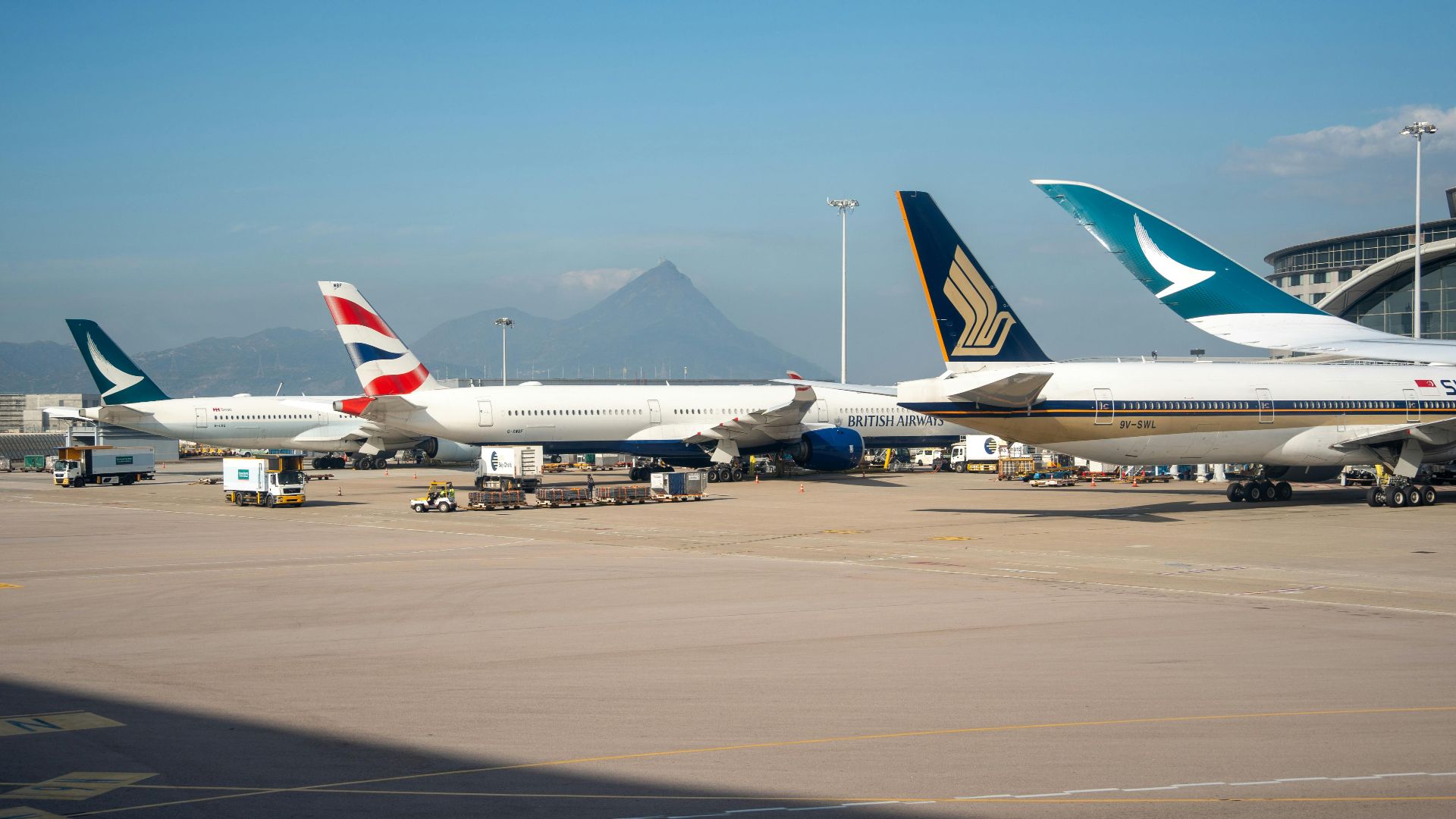 Multiple commercial airplanes parked at an airport terminal with a mountain view in the background.